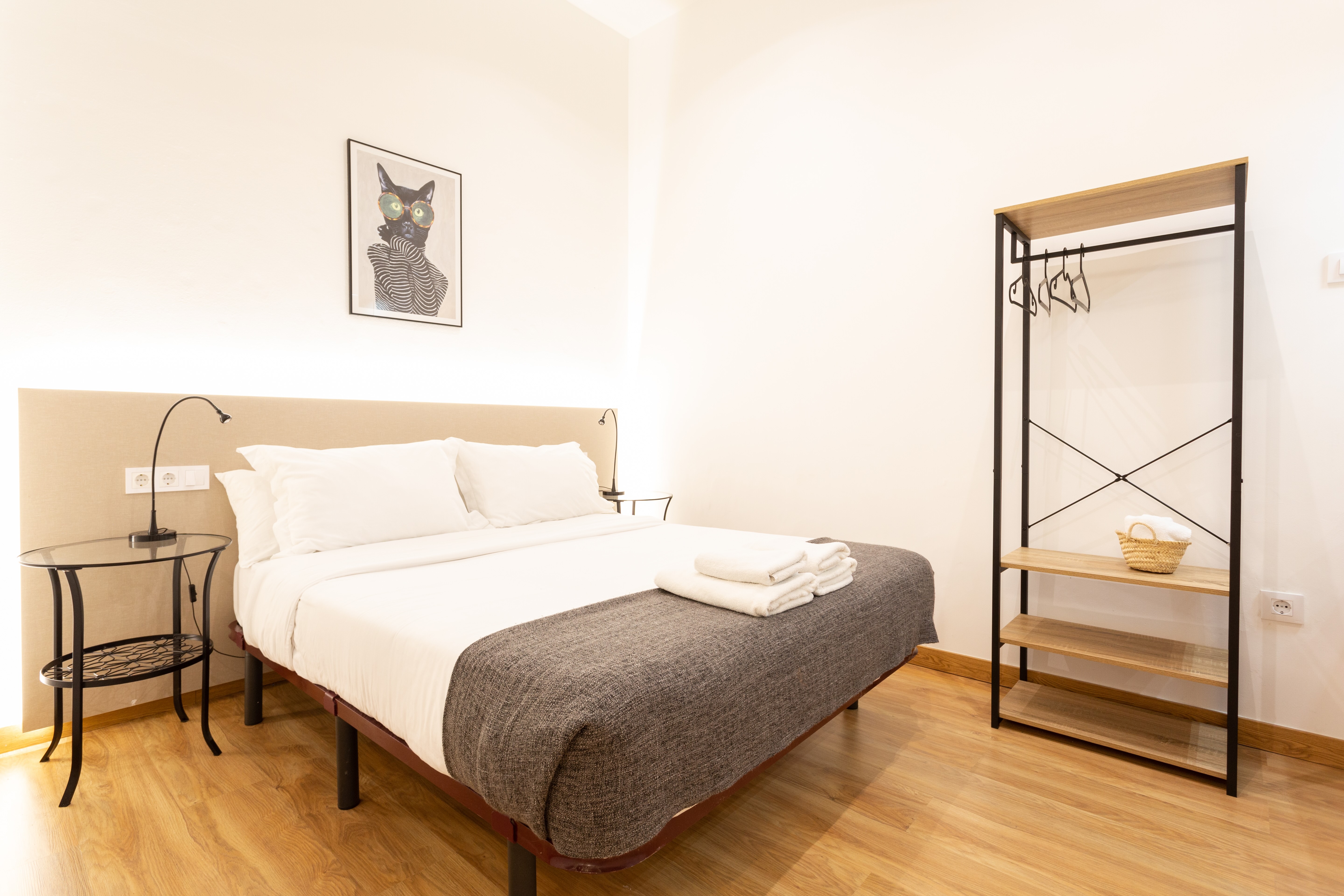 Minimalist bedroom with a double bed with white linens, gray blanket, two bedside tables with lamps, and open wooden shelving with hangers and a basket of towels.
