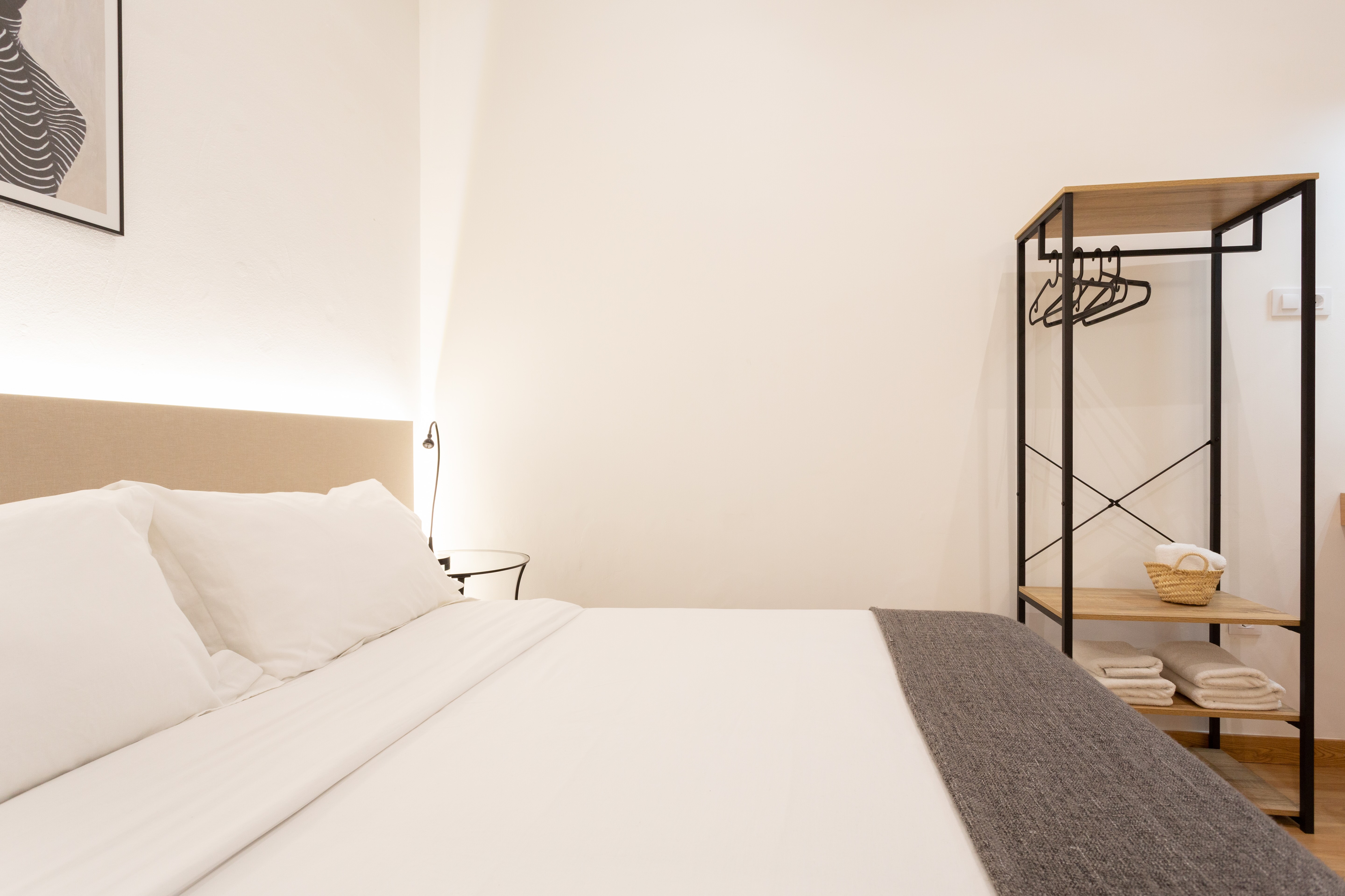 Minimalist bedroom with a neatly made bed featuring white linens and a grey throw, a bedside table with a lamp, and an open wardrobe holding hangers and folded towels.