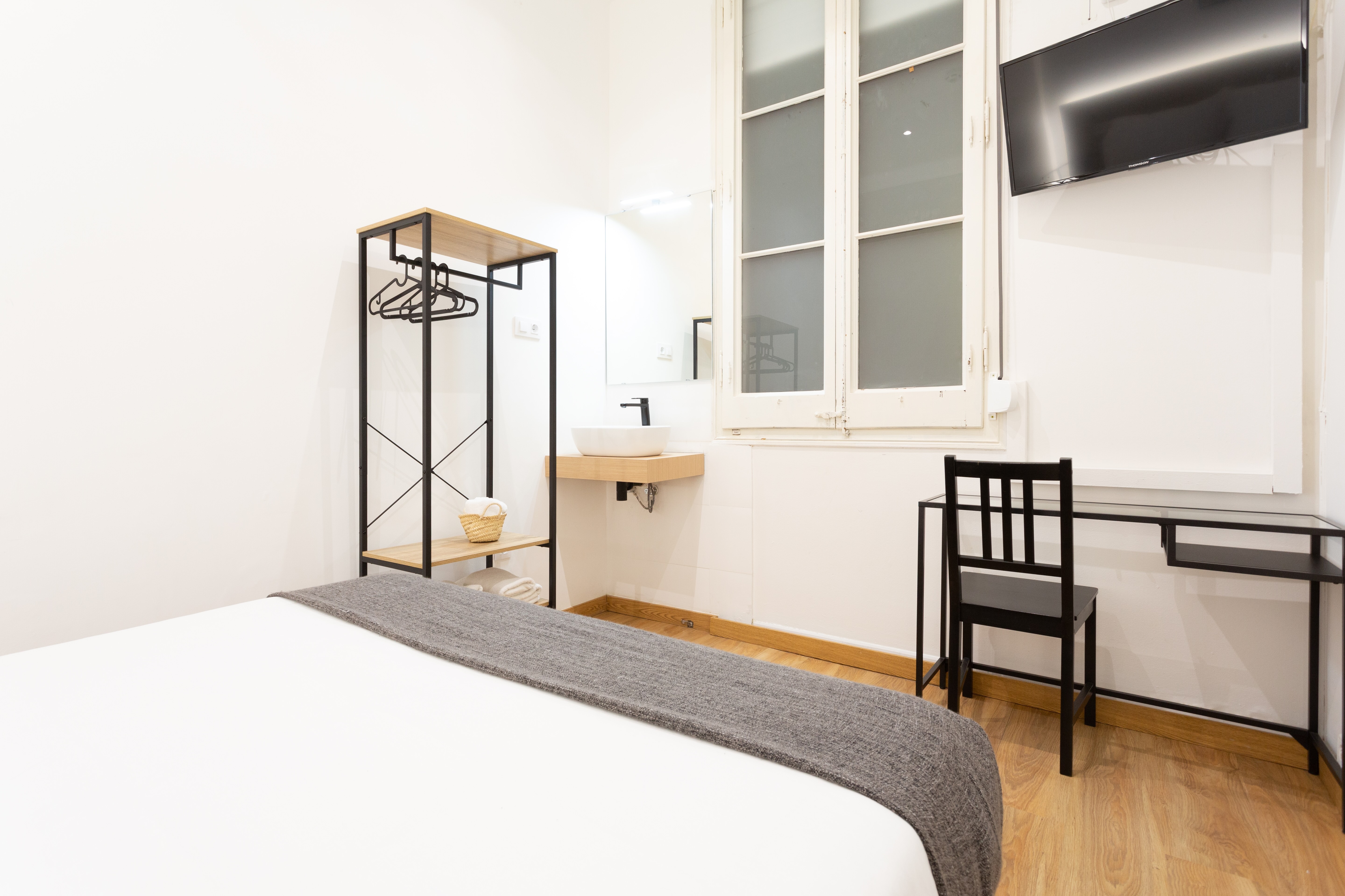 Bright minimalist bedroom with white walls, a bed with gray blanket, black metal clothes rack, wall-mounted sink, black desk and chair, and a TV mounted on the wall.