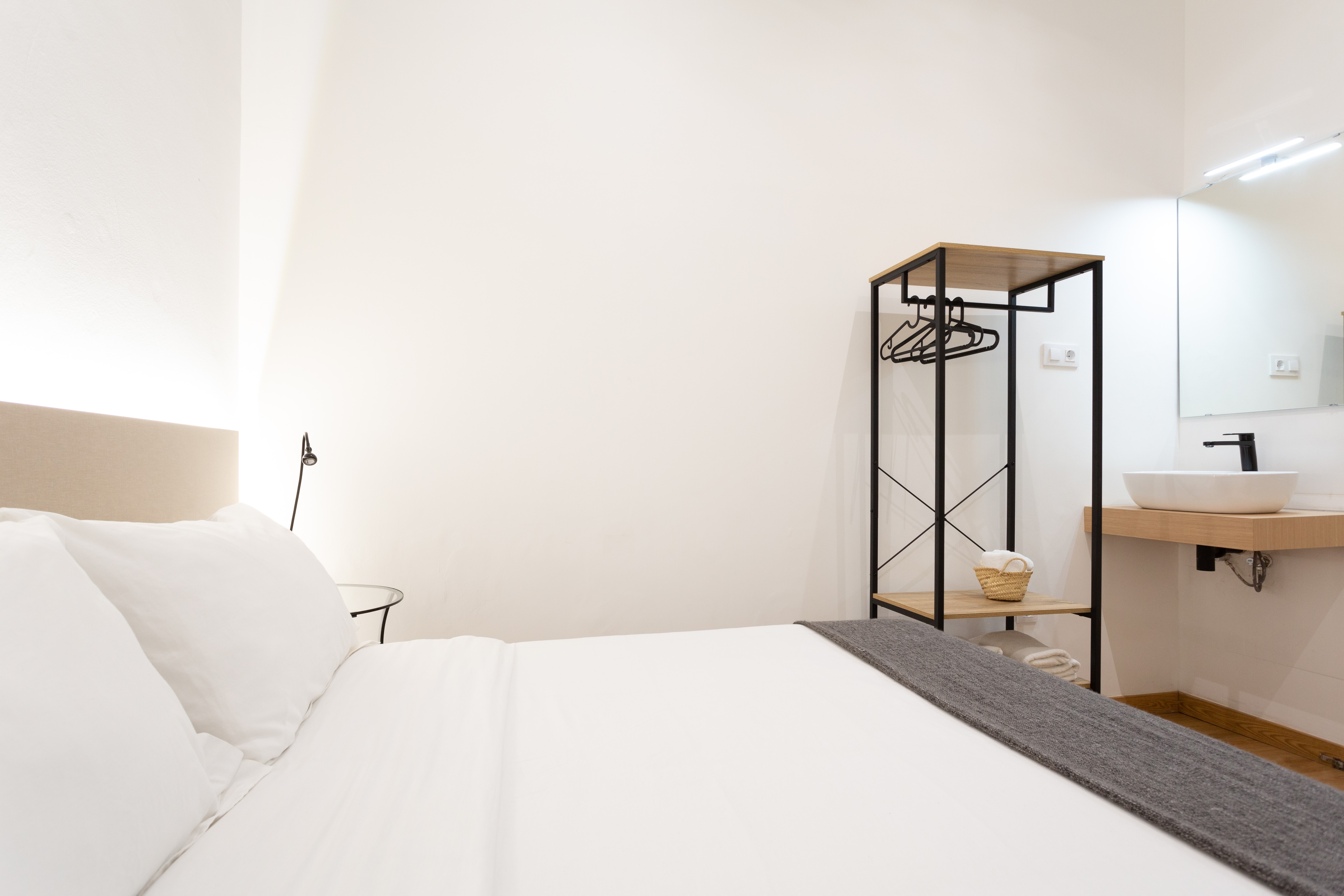 Minimalist bedroom with white bedding, a gray throw, black metal and wood open wardrobe with hangers, and a small sink with a mirror.