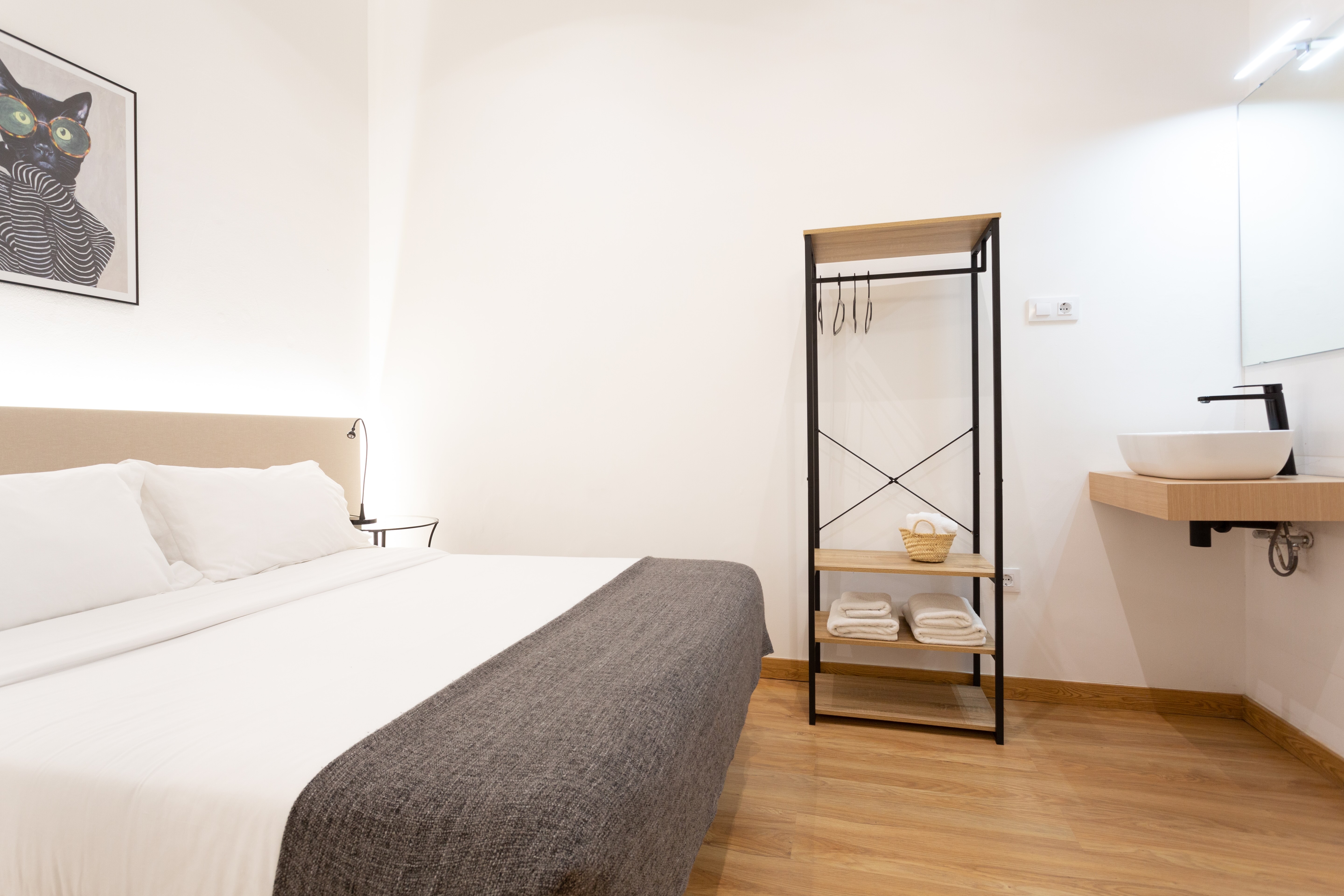 Minimalist bedroom with a large bed, gray blanket, open wooden shelf with towels, and wall-mounted sink.