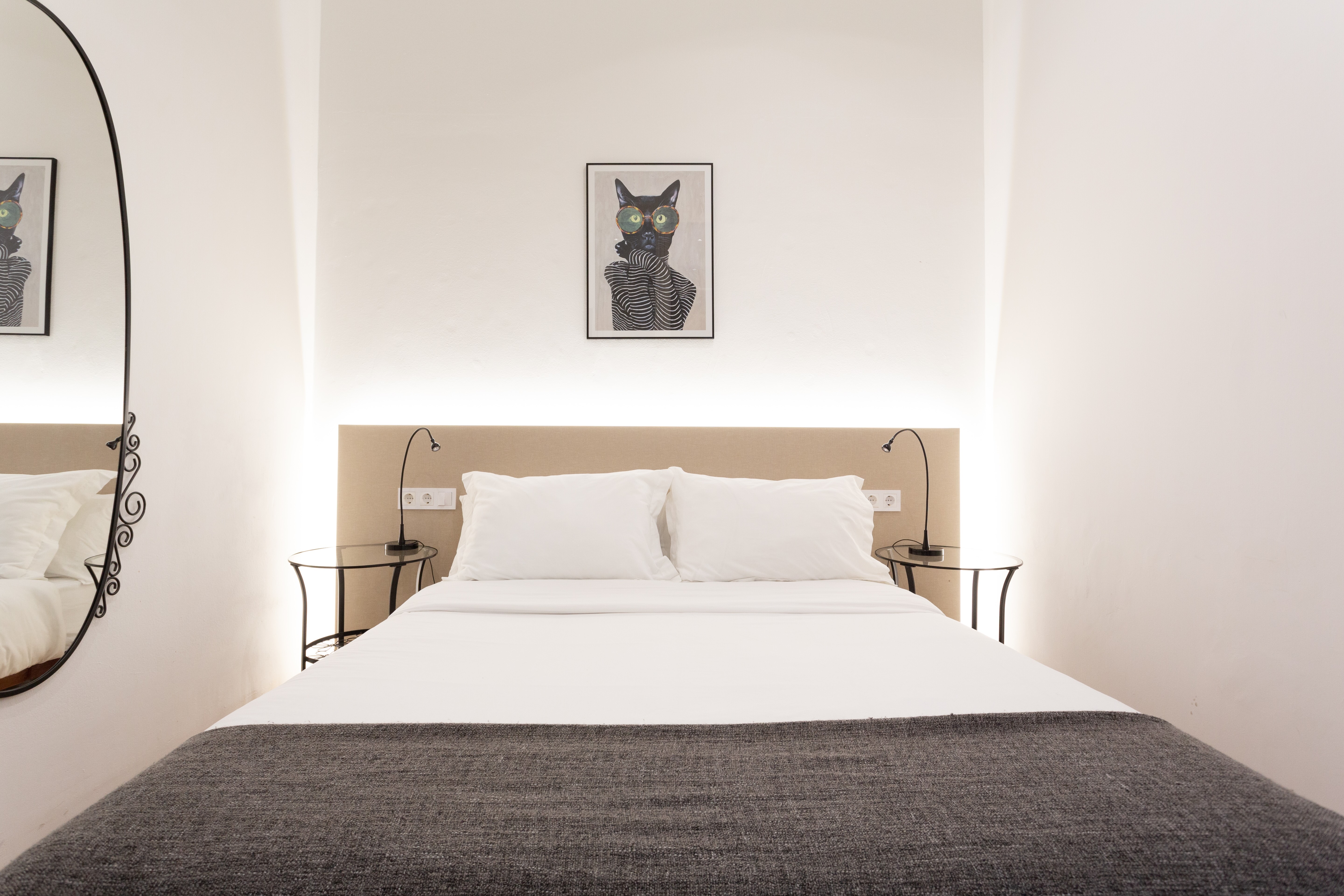 Minimalist bedroom with a neatly made double bed featuring white pillows and sheets, a gray blanket, small round bedside tables, and a framed artwork of a cat with glasses above the headboard.