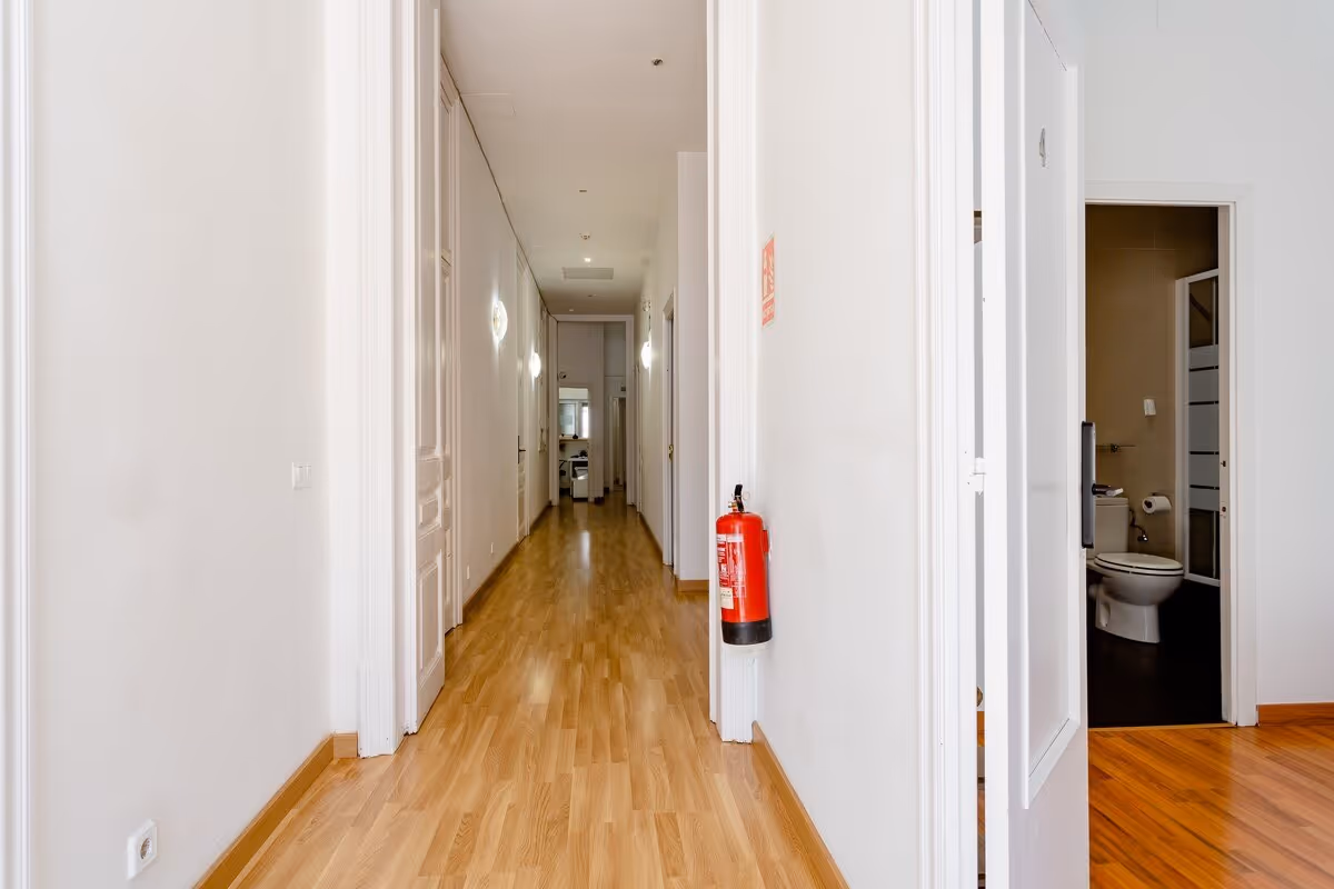 Long hallway with wooden floor, white walls, multiple doors, a fire extinguisher on the right wall, and a partially visible bathroom with a toilet.