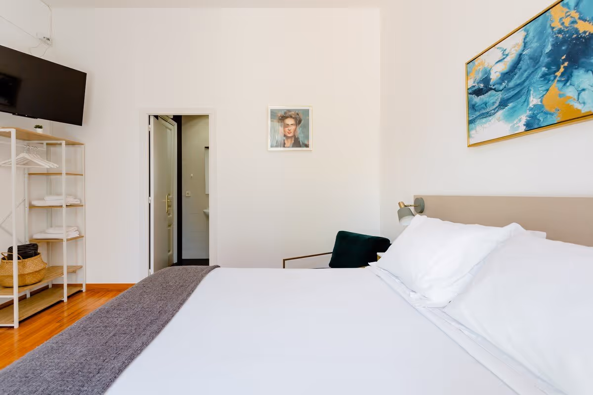 Minimalist bedroom with a large white bed, gray blanket, wall-mounted TV, open shelving with towels, and abstract blue wall art.
