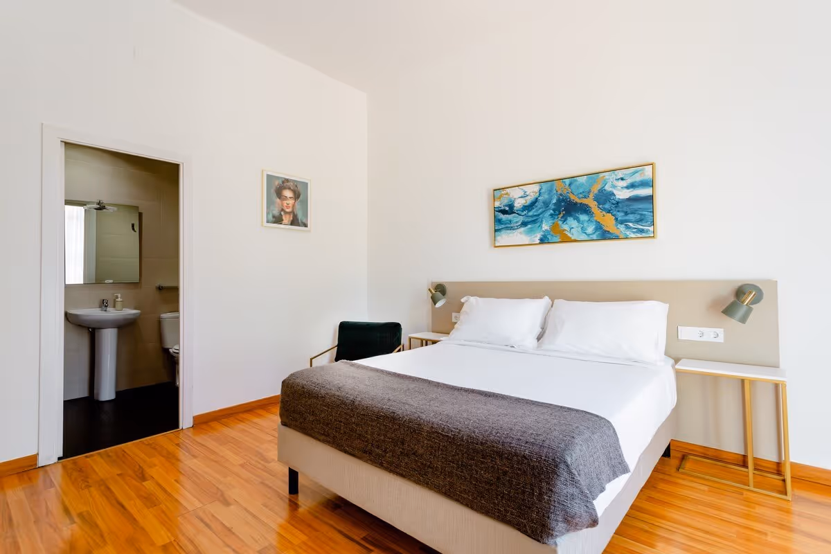 Minimalist bedroom with a double bed, white sheets, gray blanket, abstract wall art, wooden floor, and an open door to a bathroom.