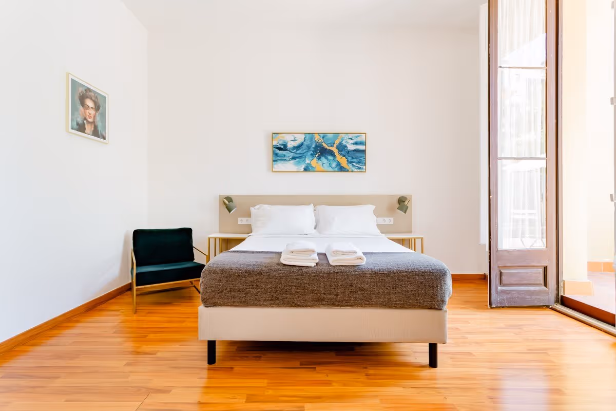 Minimalist bedroom with a double bed covered in white linens and gray blanket, two folded towels on top, a black armchair to the left, abstract blue and gold painting above the headboard, and a tall wooden door on the right letting in natural light.
