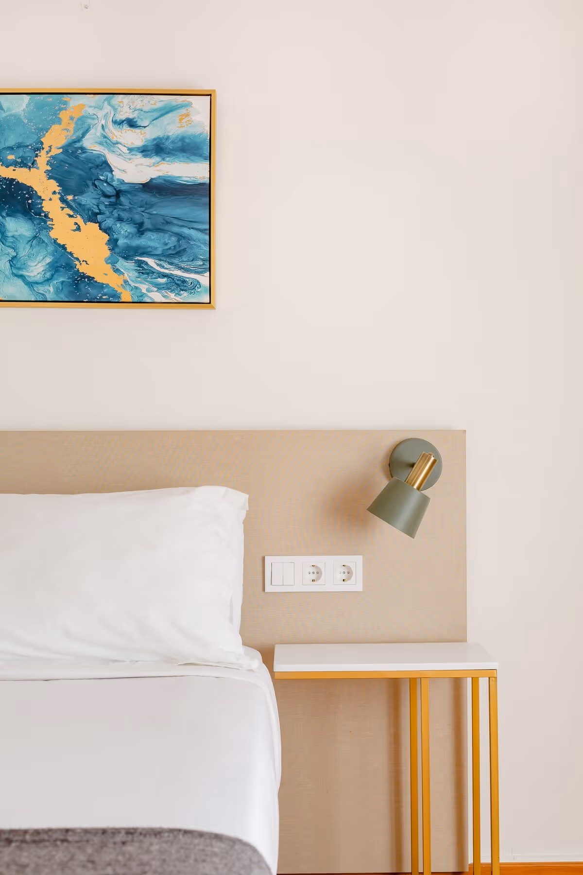 Minimalist bedroom corner with white bed and pillow, beige headboard, small white and yellow nightstand, green wall lamp, and abstract blue and gold painting.