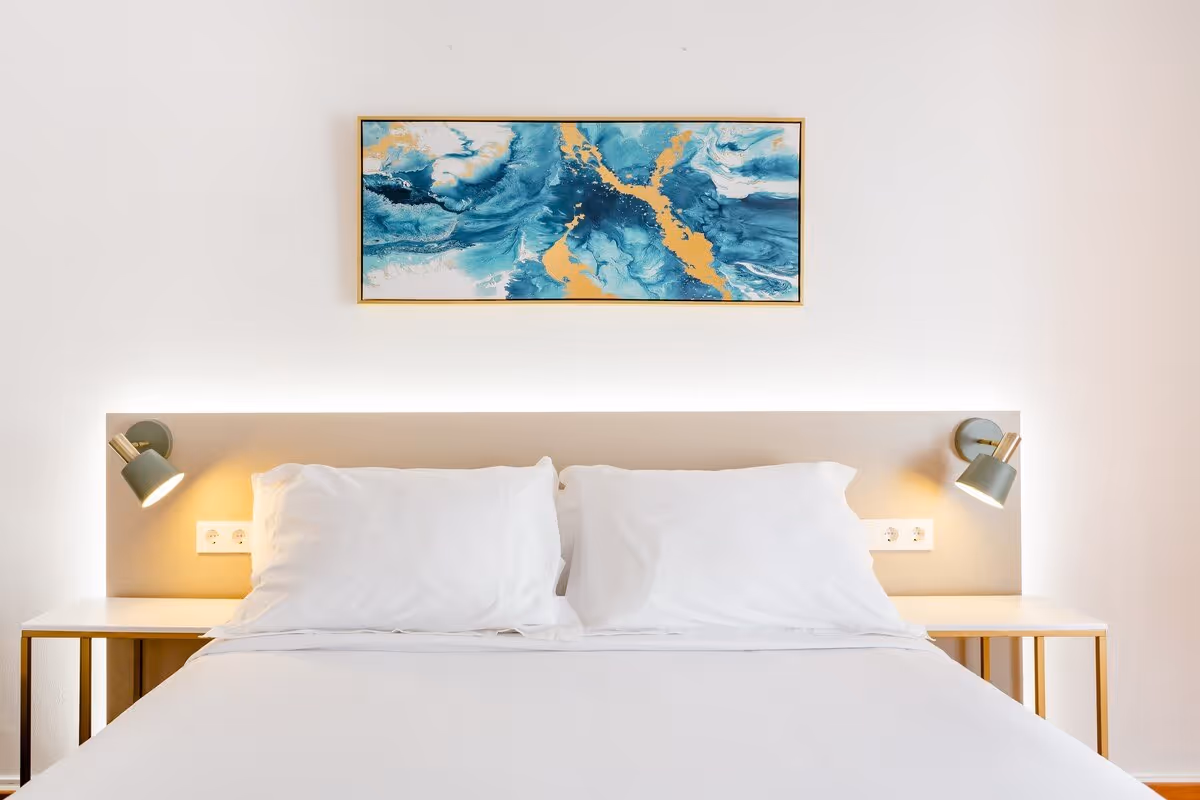 Minimalist bedroom with a white bed, two white pillows, bedside tables, wall lamps, and blue abstract painting above the headboard.