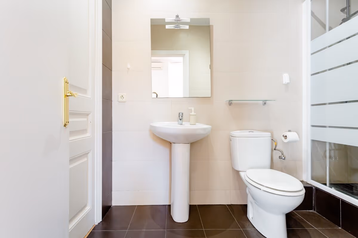 Modern bathroom with white pedestal sink, wall mirror, toilet, and glass shower door on brown tiled floor.