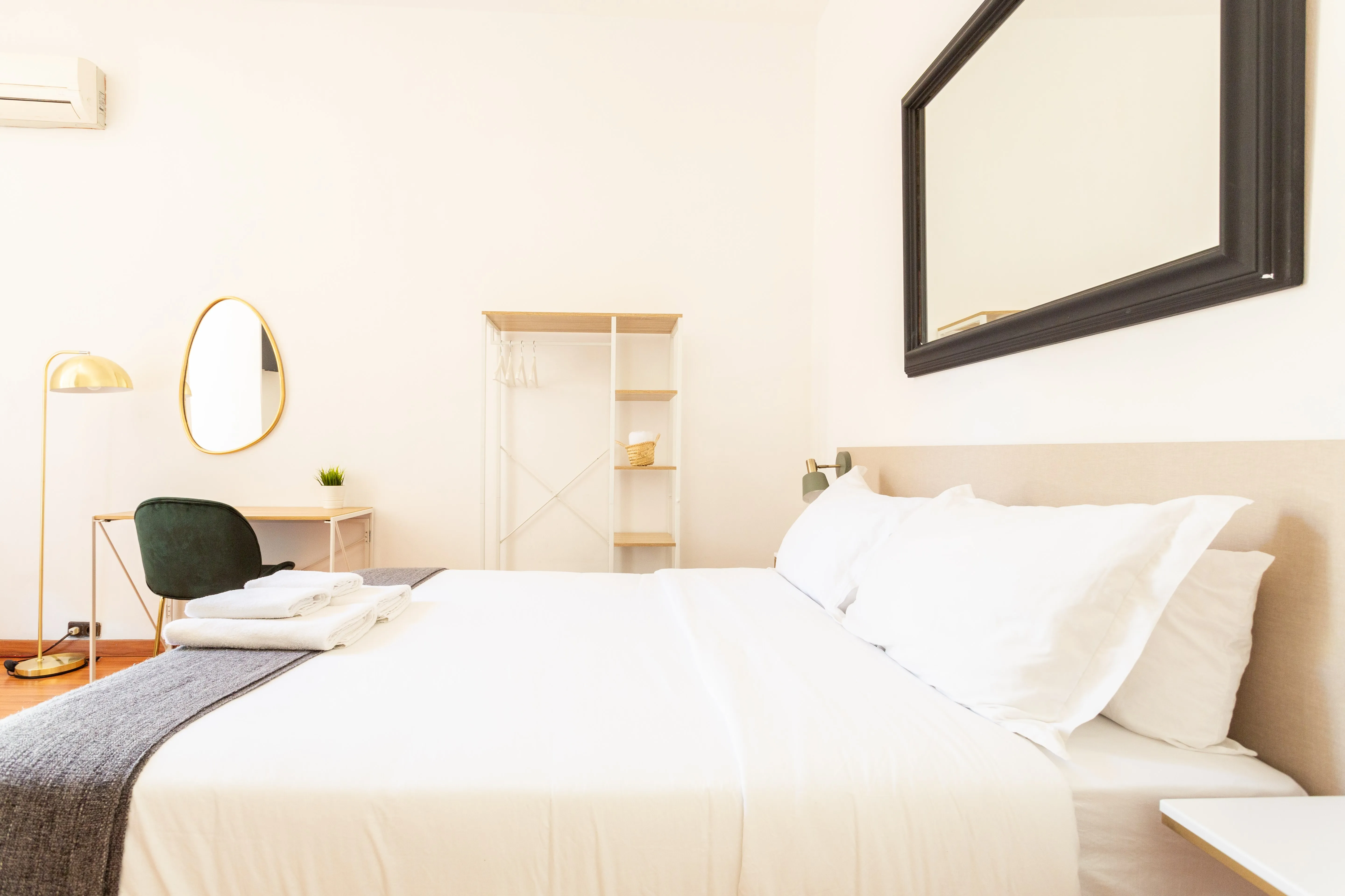 Bright minimalist bedroom with a large white bed, gray throw, folded towels, green chair, desk, floor lamp, wall mirror, and open shelving unit.