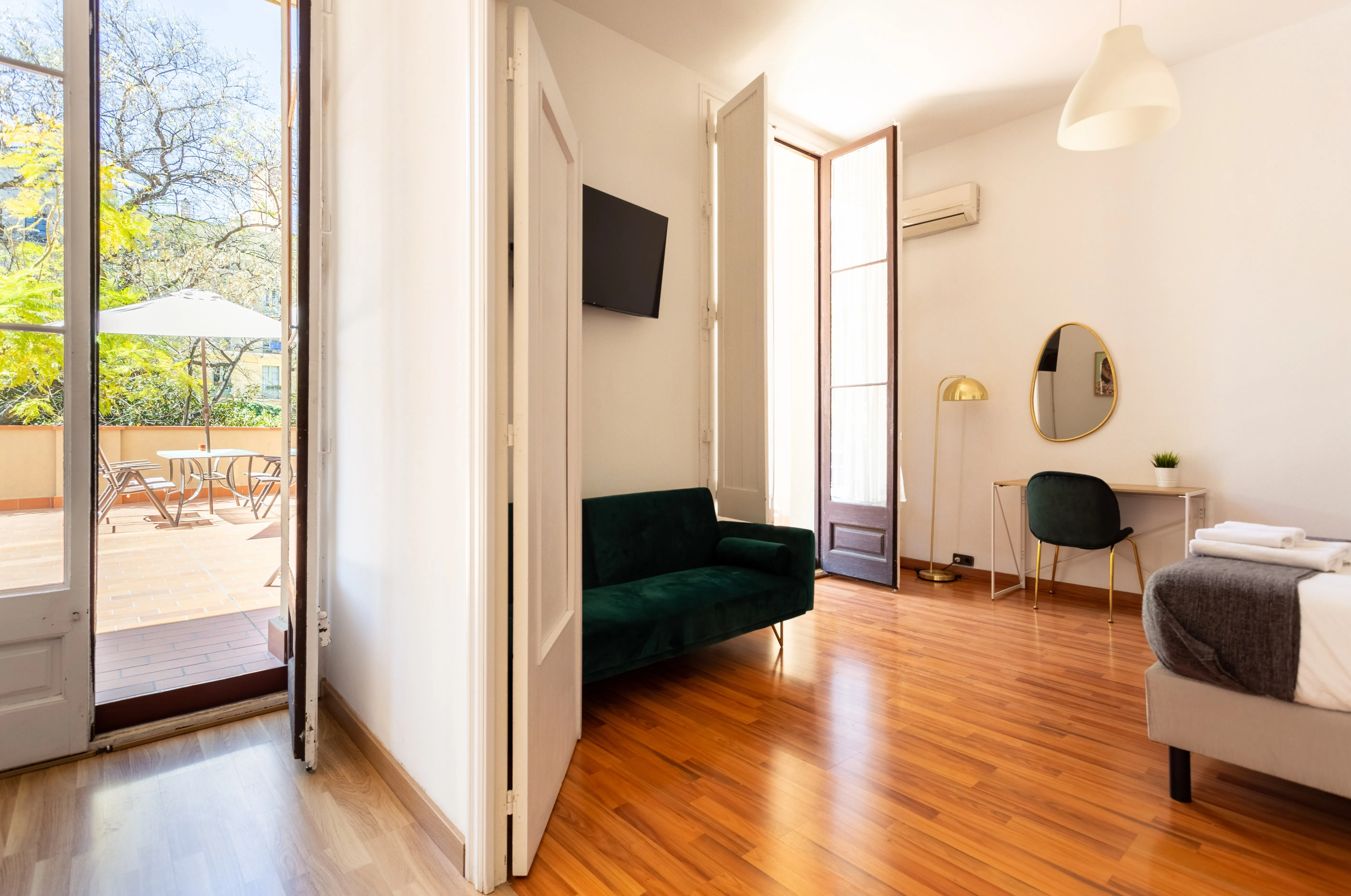 Bright room with wooden floor, green velvet sofa, desk with round mirror and chair, bed corner, and open glass doors leading to a sunlit terrace with outdoor furniture and umbrella.