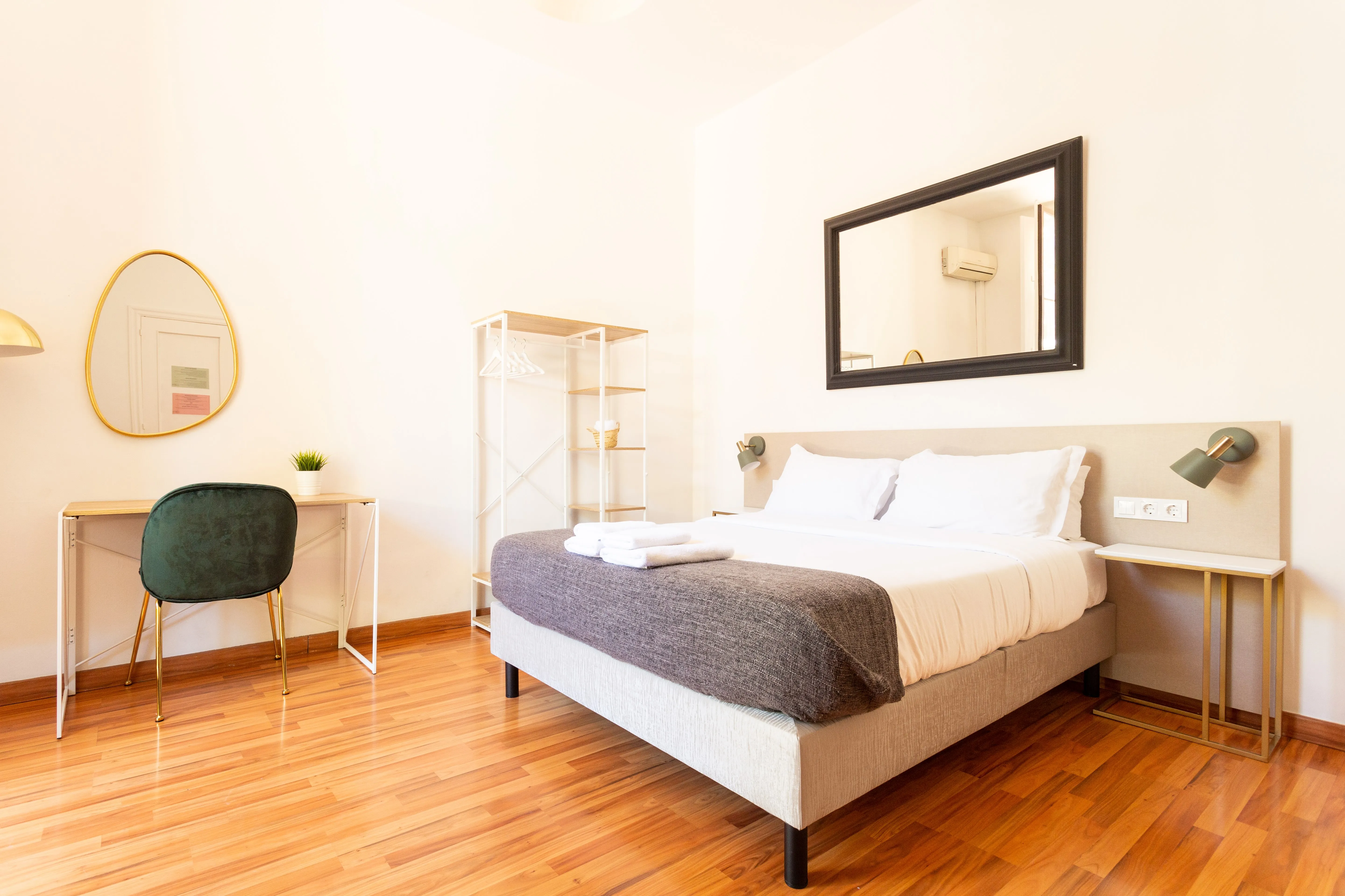 Minimalist bedroom with a bed, white sheets, gray blanket, bedside table, mirror, desk with green chair, and wooden floor.
