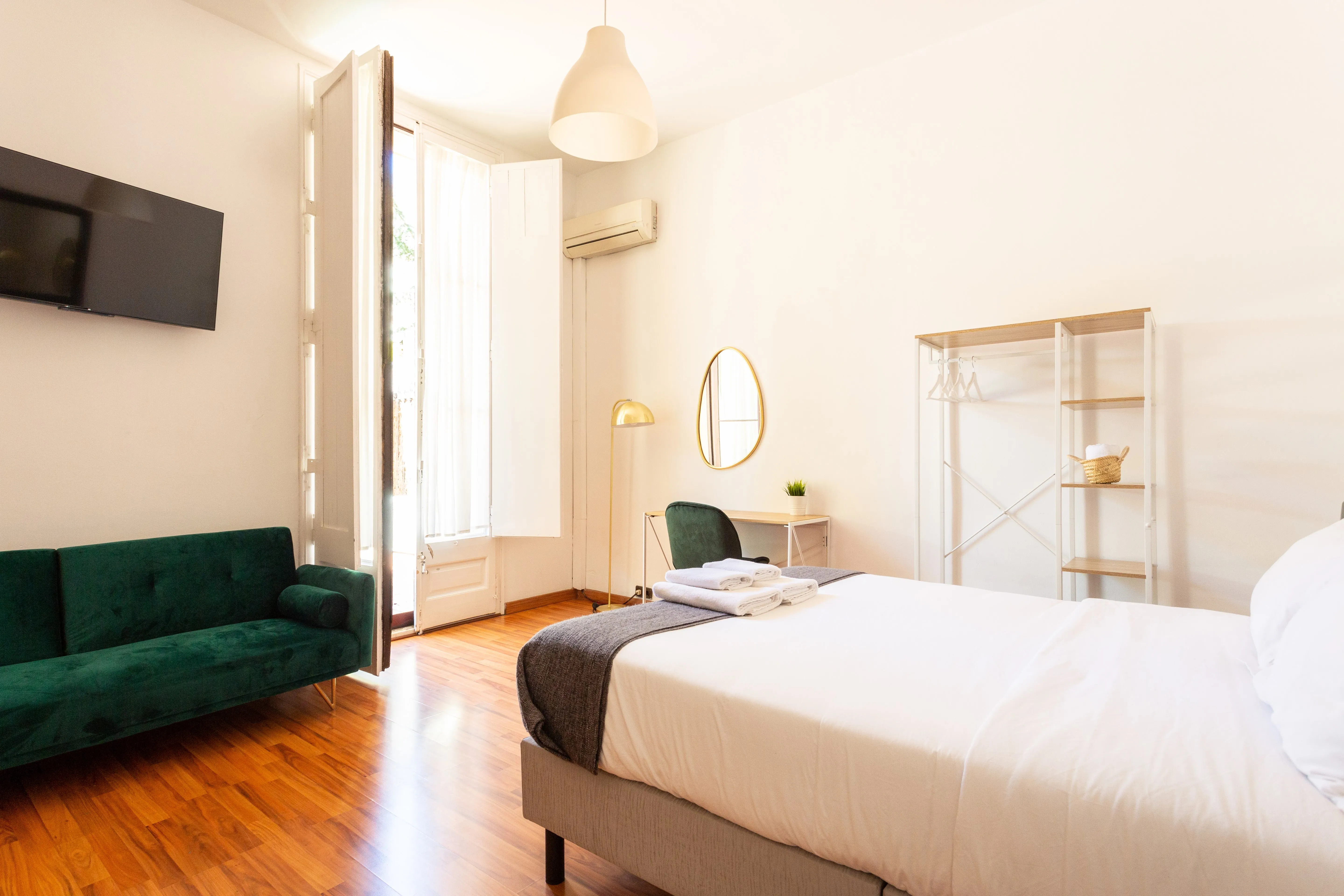 Bright bedroom with white walls, wooden floor, green velvet sofa, large bed with white linens, and open window with sheer curtains.