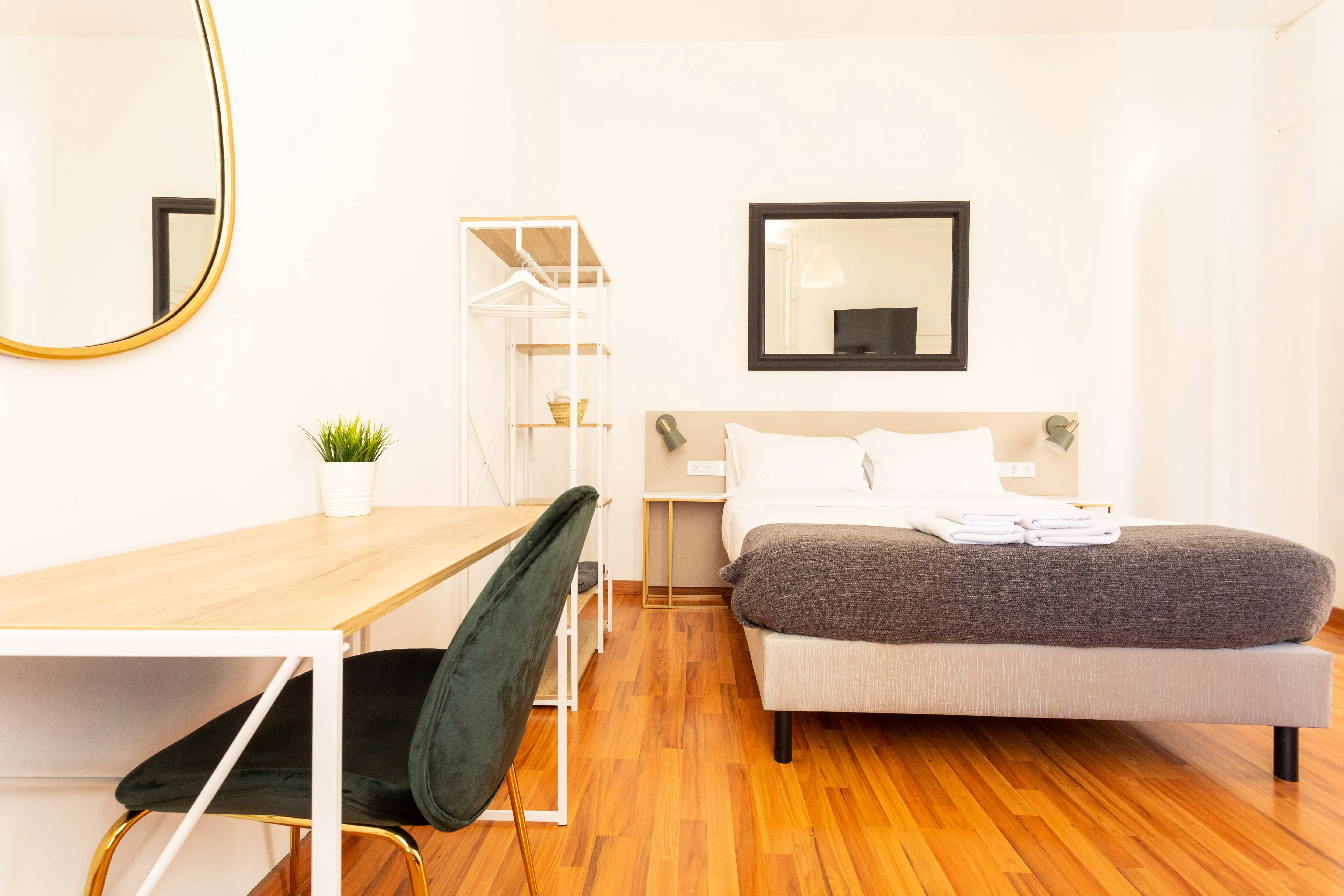 Minimalist bedroom with a double bed, dark gray blanket, white pillows, a wooden desk with a green chair, and a large mirror on the wall.
