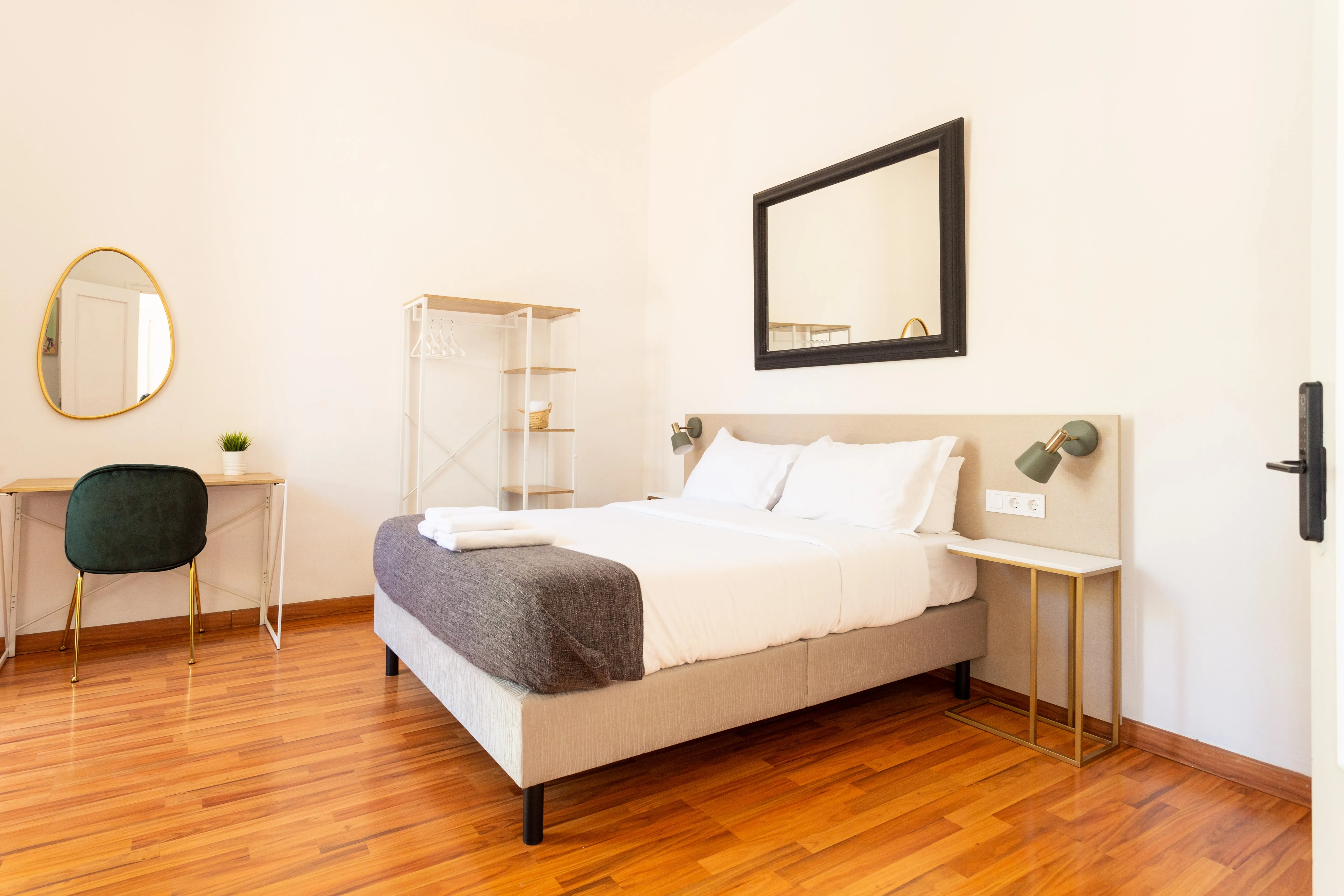 Minimalist bedroom with a double bed featuring white linens and gray blanket, wooden floor, small desk with green chair, and wall-mounted mirror.