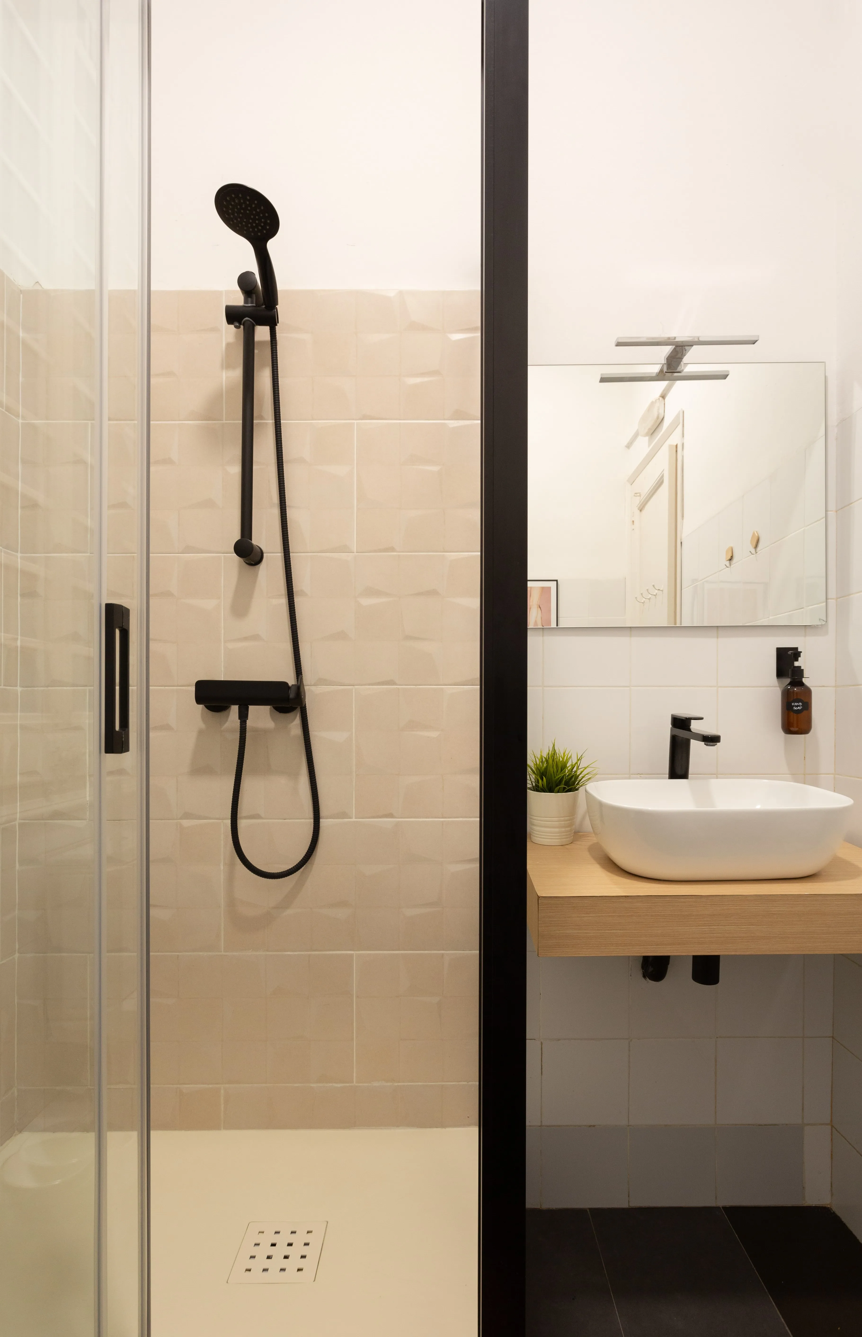 Modern bathroom with beige tiled shower featuring a black handheld showerhead and a wooden floating vanity with a white vessel sink and black faucet.