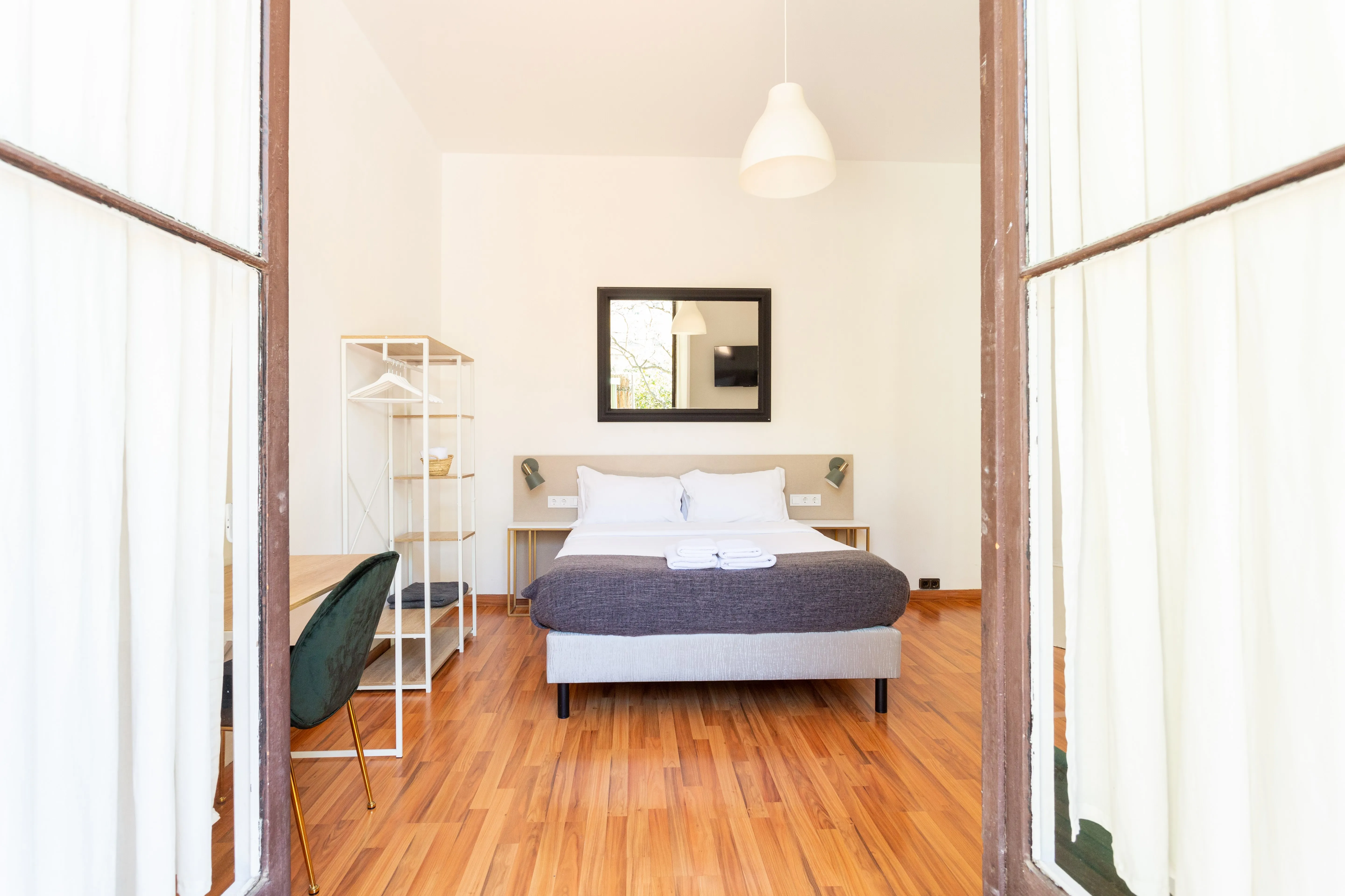 Bright bedroom with wooden floor, double bed with white and gray bedding, a mirror above headboard, a small open wardrobe, and a dark green chair at a desk.