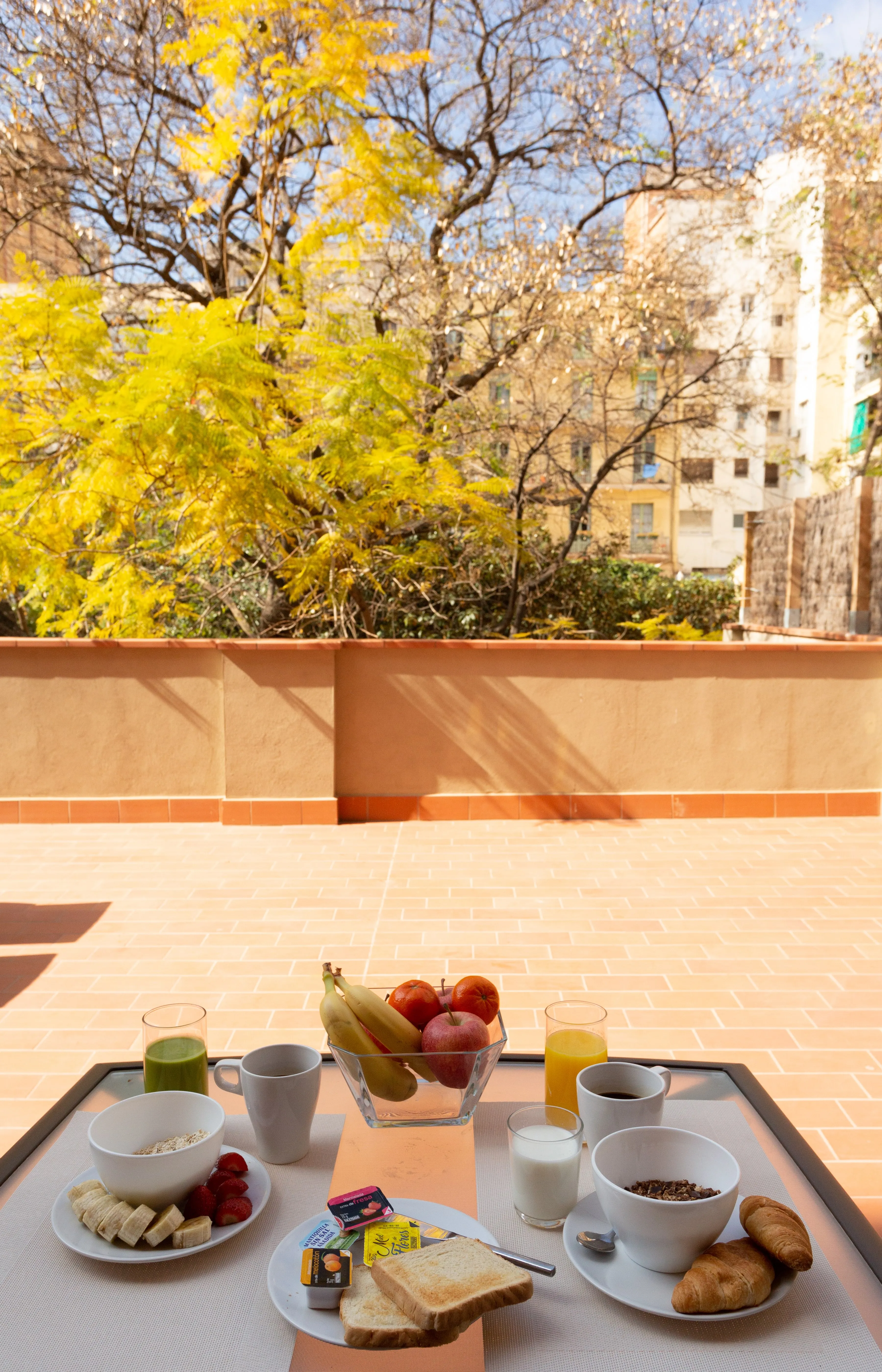 Outdoor breakfast setup with bowls of cereal and fruit, toast with spreads, croissants, and glasses of juice and milk on a table.
