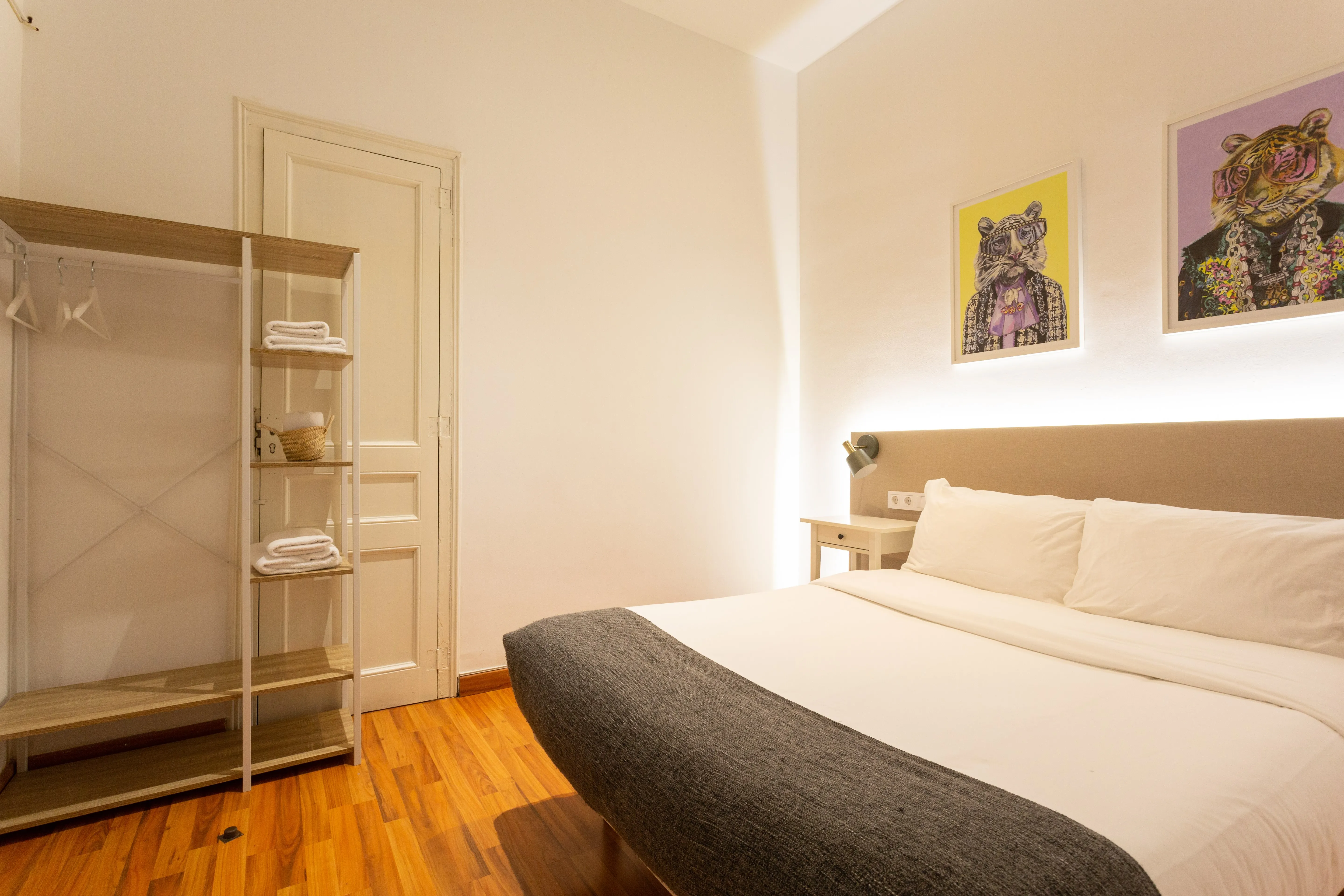 Minimalist bedroom with a large bed, white bedding, gray throw blanket, wooden floor, and wall art of tigers wearing glasses.