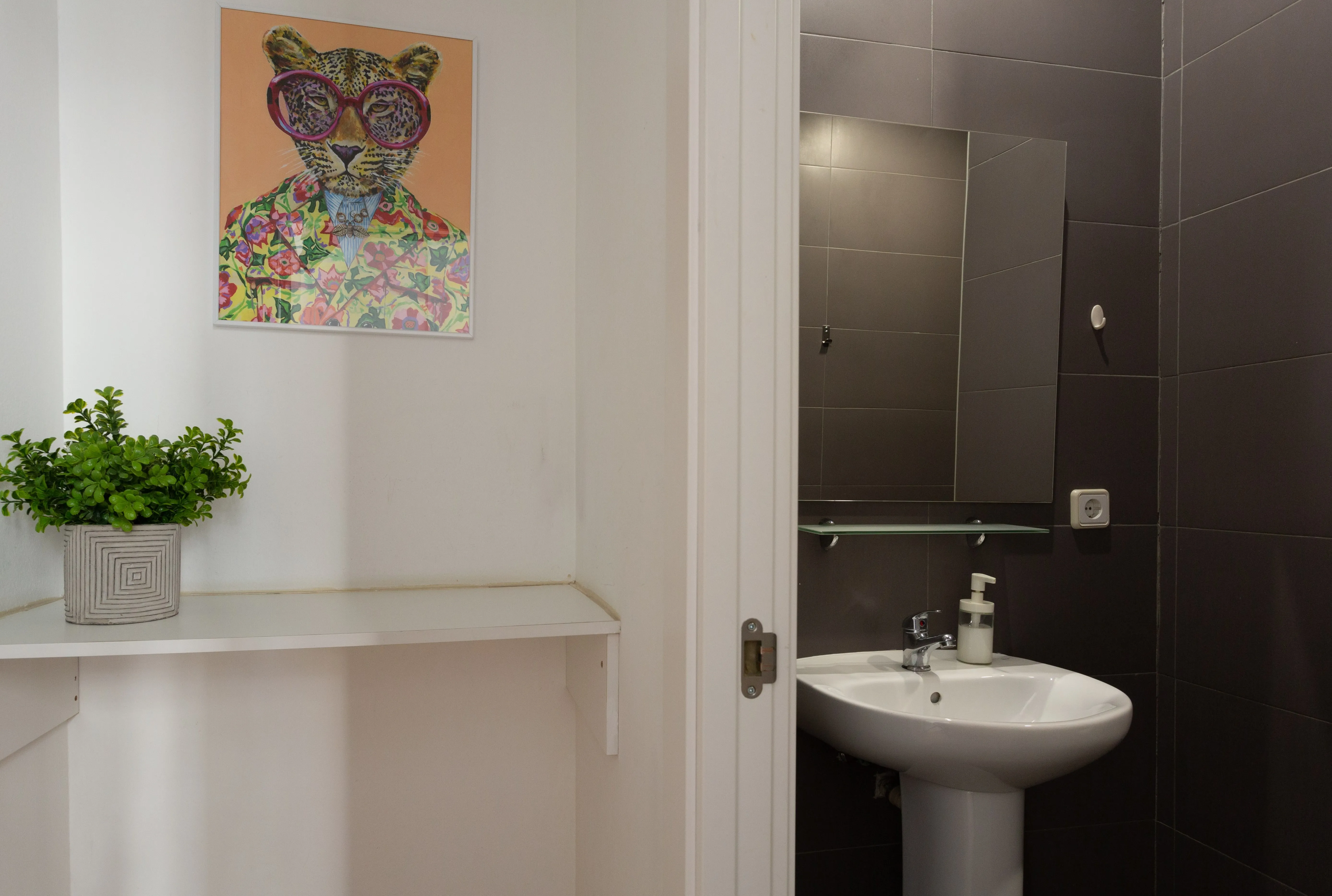 White shelf with a green potted plant and a colorful artwork of a leopard wearing glasses, next to a bathroom with a white pedestal sink and a mirror on dark tiled walls.