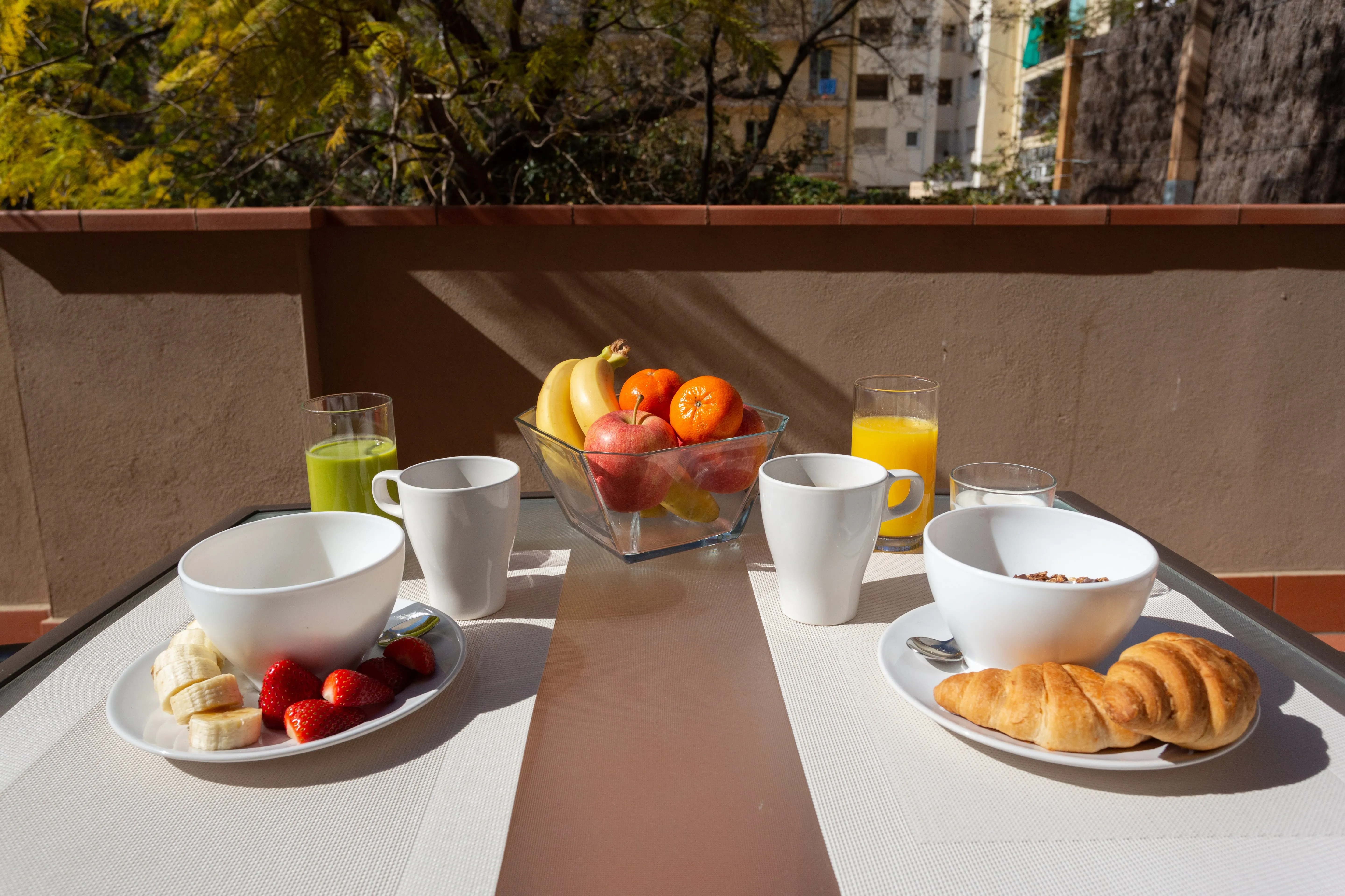 Outdoor table set with two breakfast plates, one with sliced banana and strawberries, the other with croissants and cereal bowls, alongside cups, a glass of green juice, a glass of orange juice, and a bowl of mixed fruit.