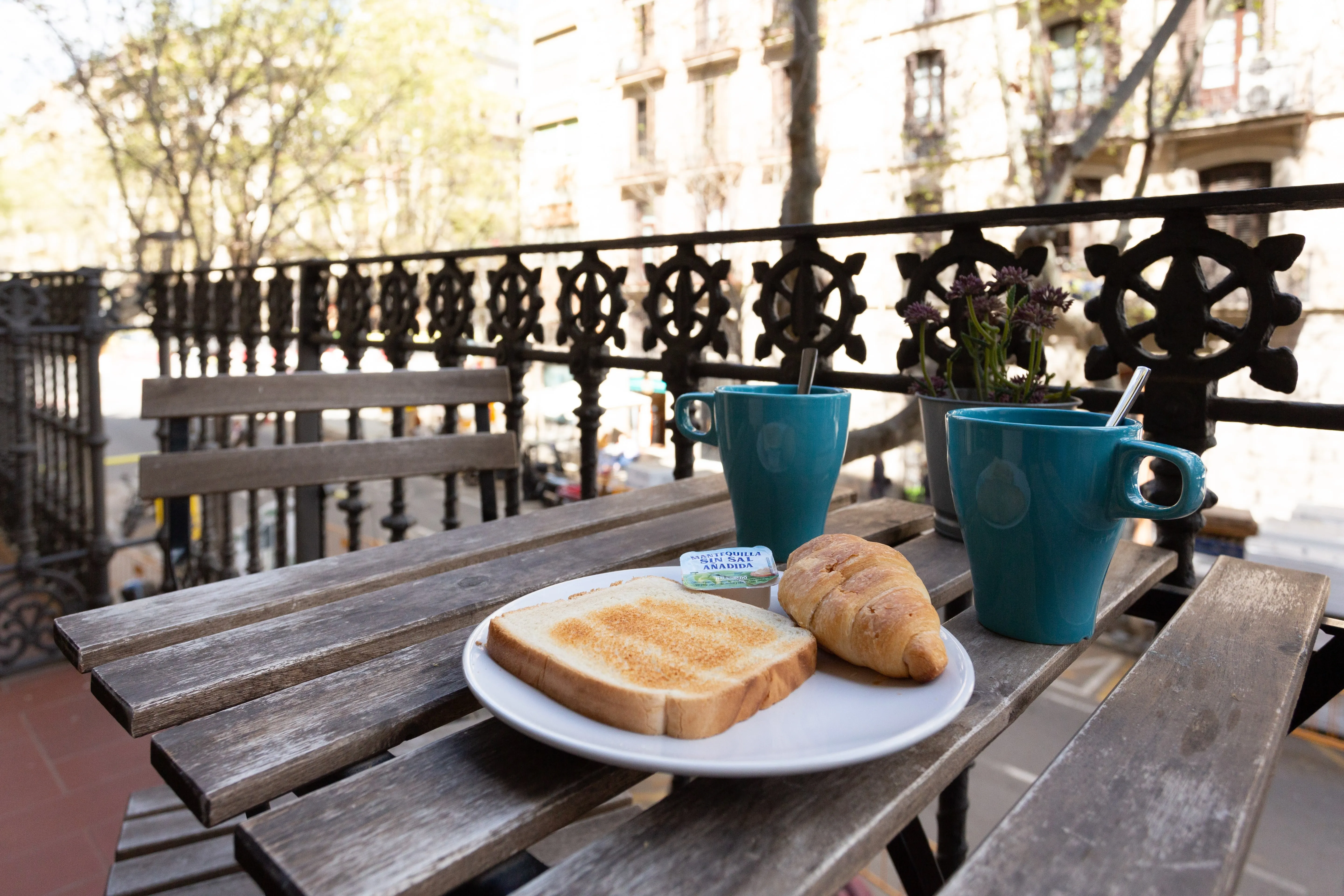 Wooden balcony table with two blue mugs, a white plate holding a slice of toasted bread, a croissant, and a packet of butter with a small potted plant in the background.