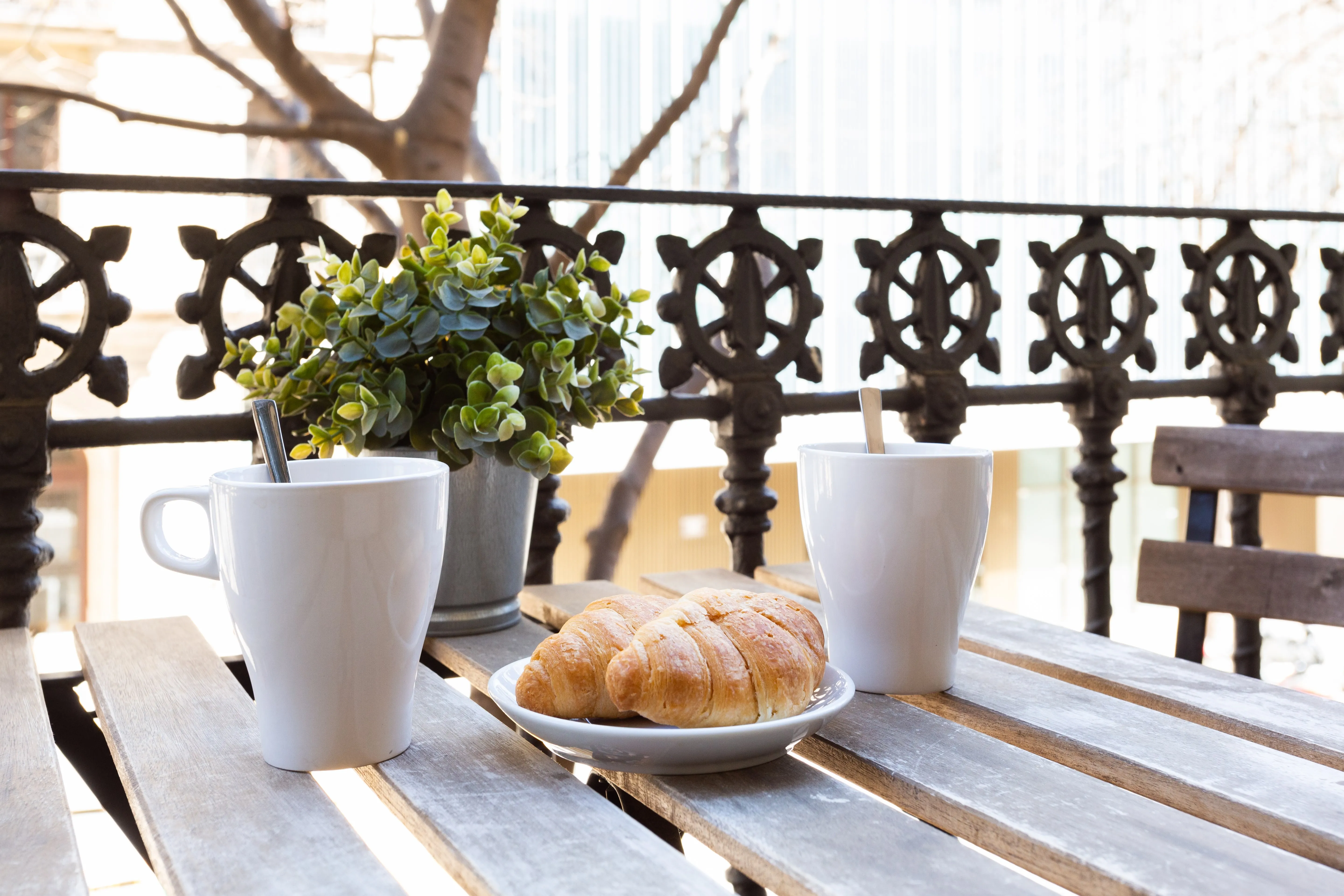 Two white mugs with spoons, a plate with two croissants, and a potted plant on a wooden outdoor table with a decorative wrought iron railing in the background.