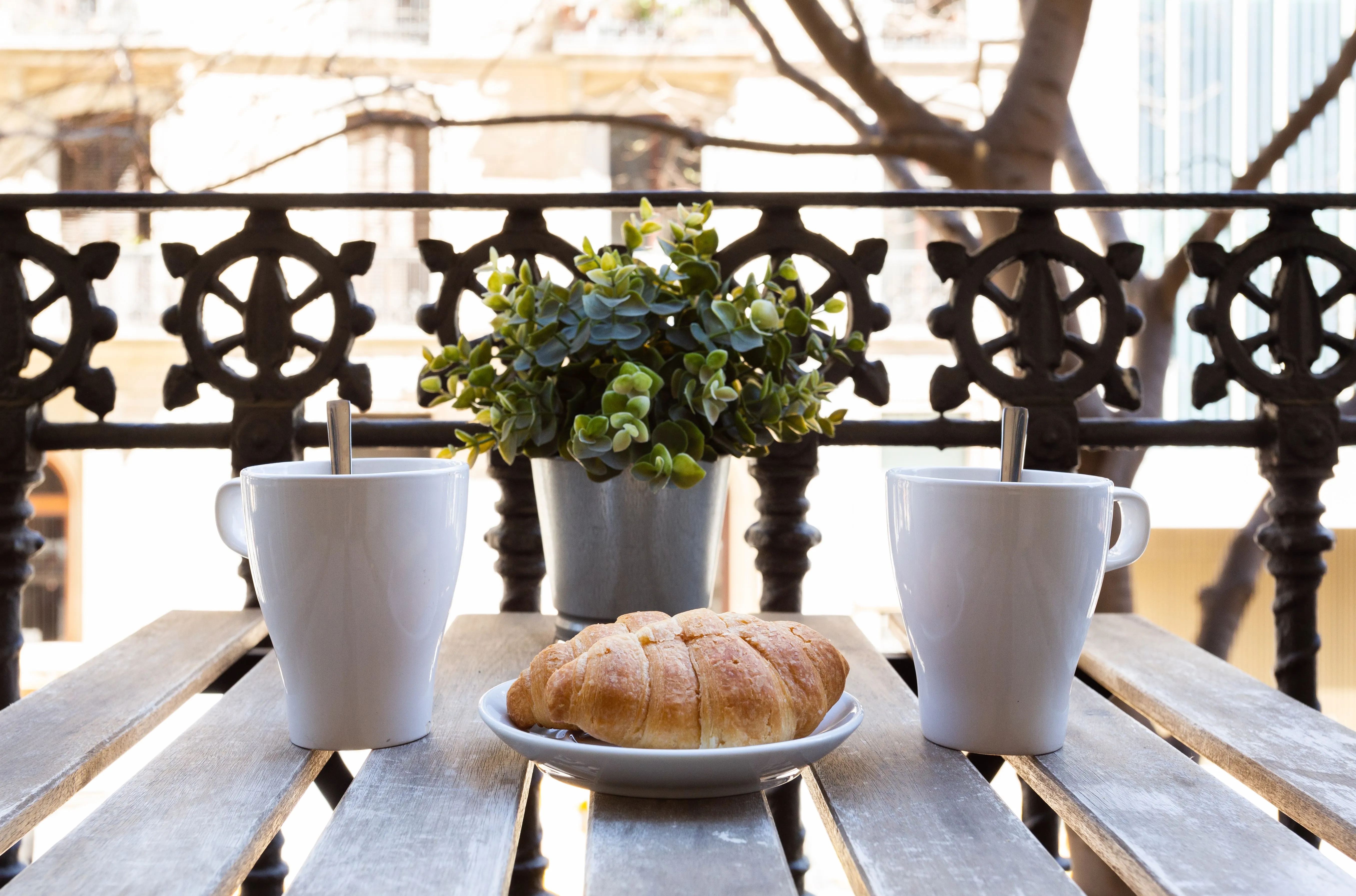 Two white mugs with spoons, a croissant on a white plate, and a potted plant on a wooden table on a balcony with decorative railing.