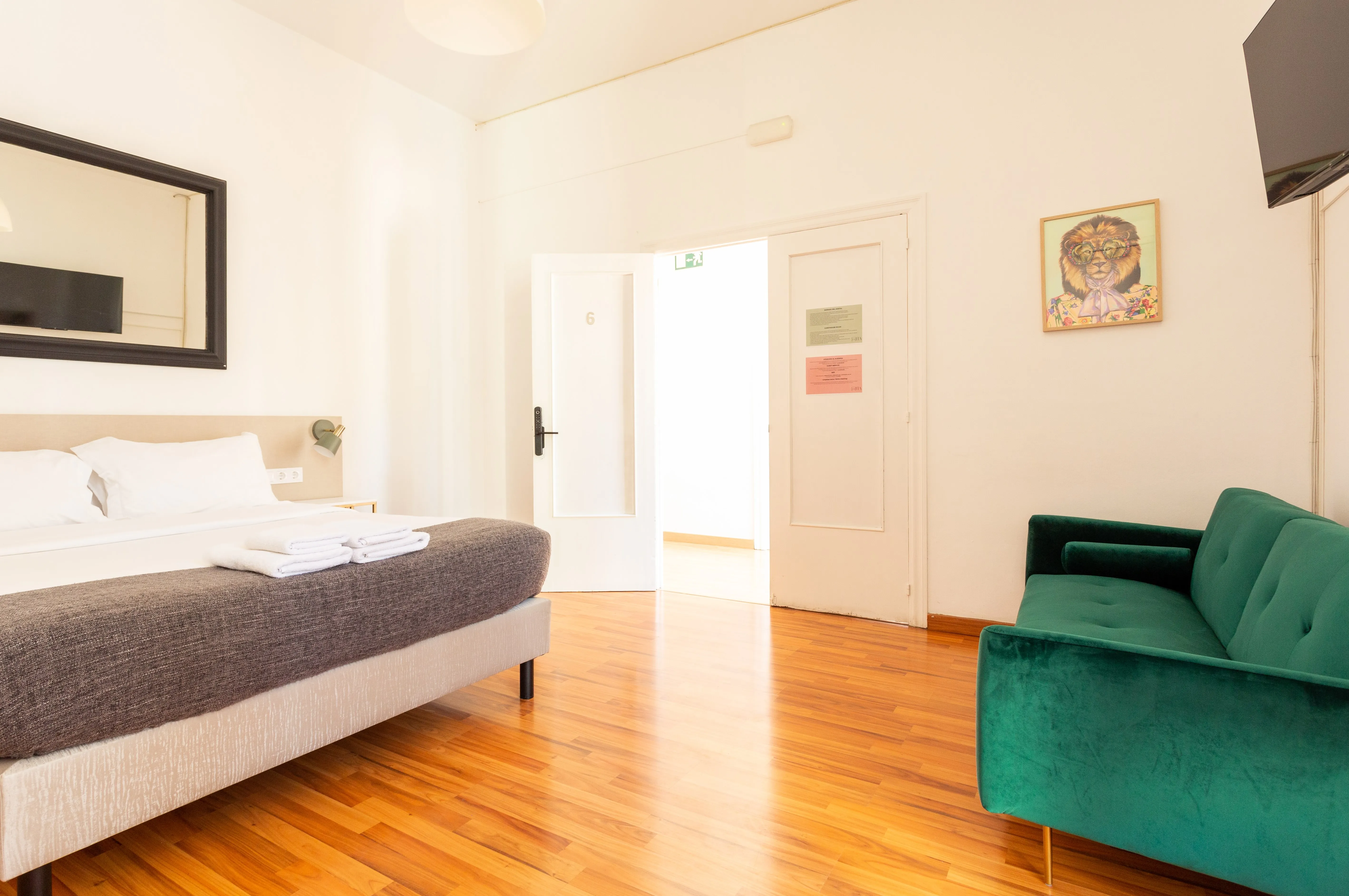 Bright hotel room with wooden floor, beige bed with gray blanket and folded towels, green sofa, wall-mounted TV, and a painting of a lion wearing glasses.