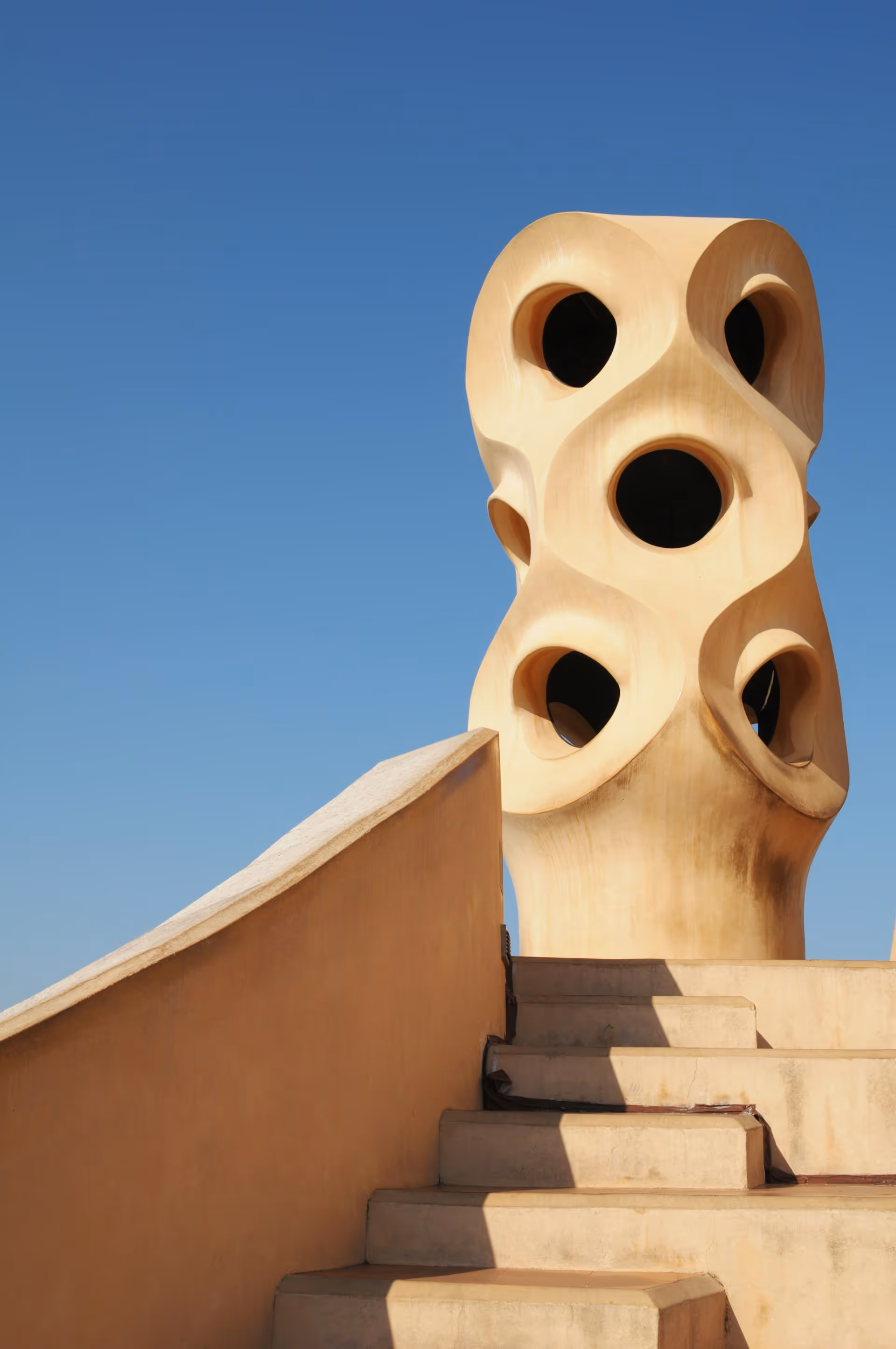 Curved beige stone stairs leading up to a unique sculptural chimney with several circular openings against a clear blue sky.