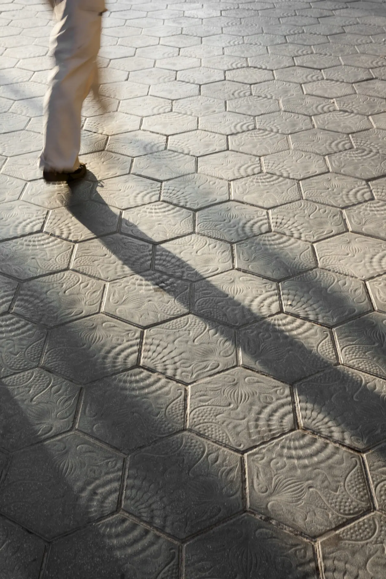 Person walking on hexagonal patterned pavement tiles casting a long shadow.