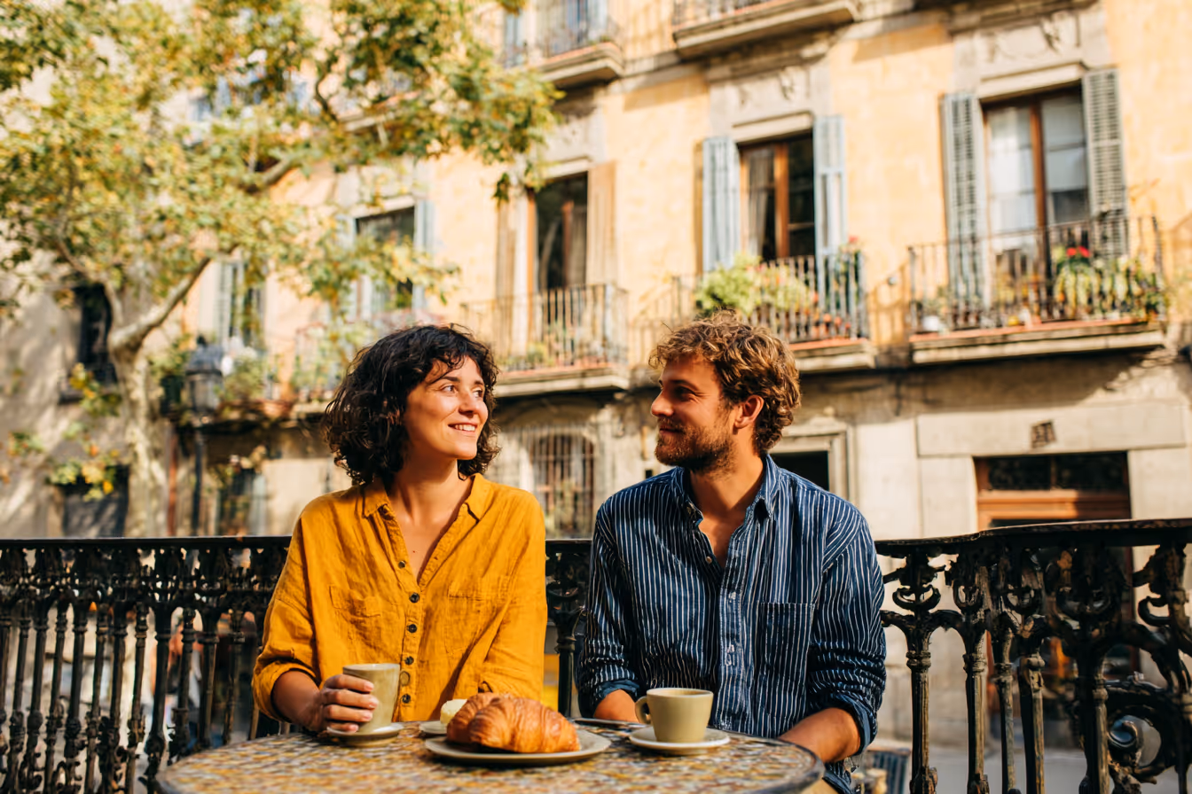 A man and woman sitting at an outdoor table with coffee cups and a croissant, smiling at each other on a sunny day.