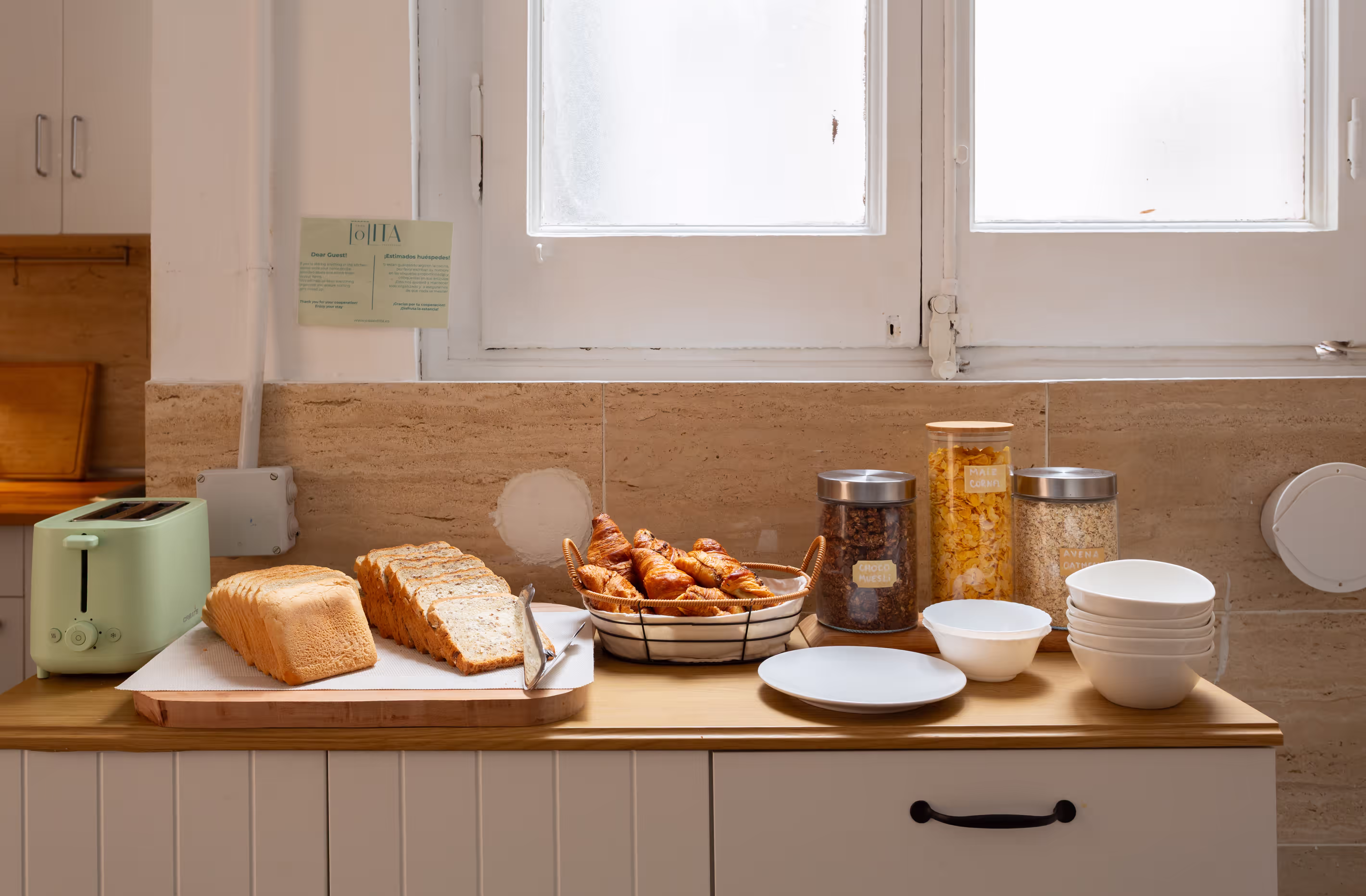 Kitchen counter with toaster, sliced bread, croissants, cereal jars, bowls, and plates under a window.