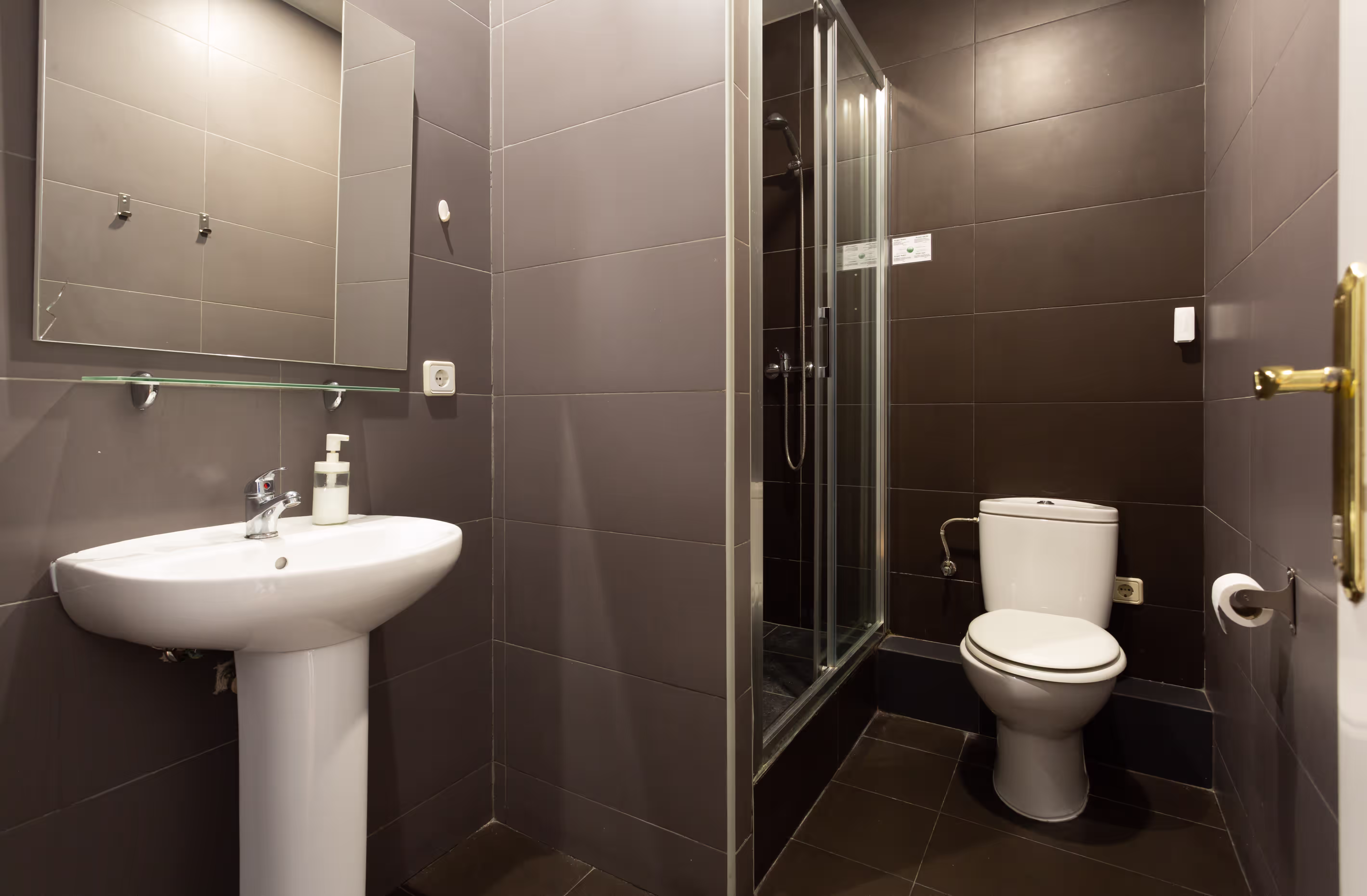 Modern bathroom with white sink, mirror, glass shower, and white toilet with dark tiled walls and floor.
