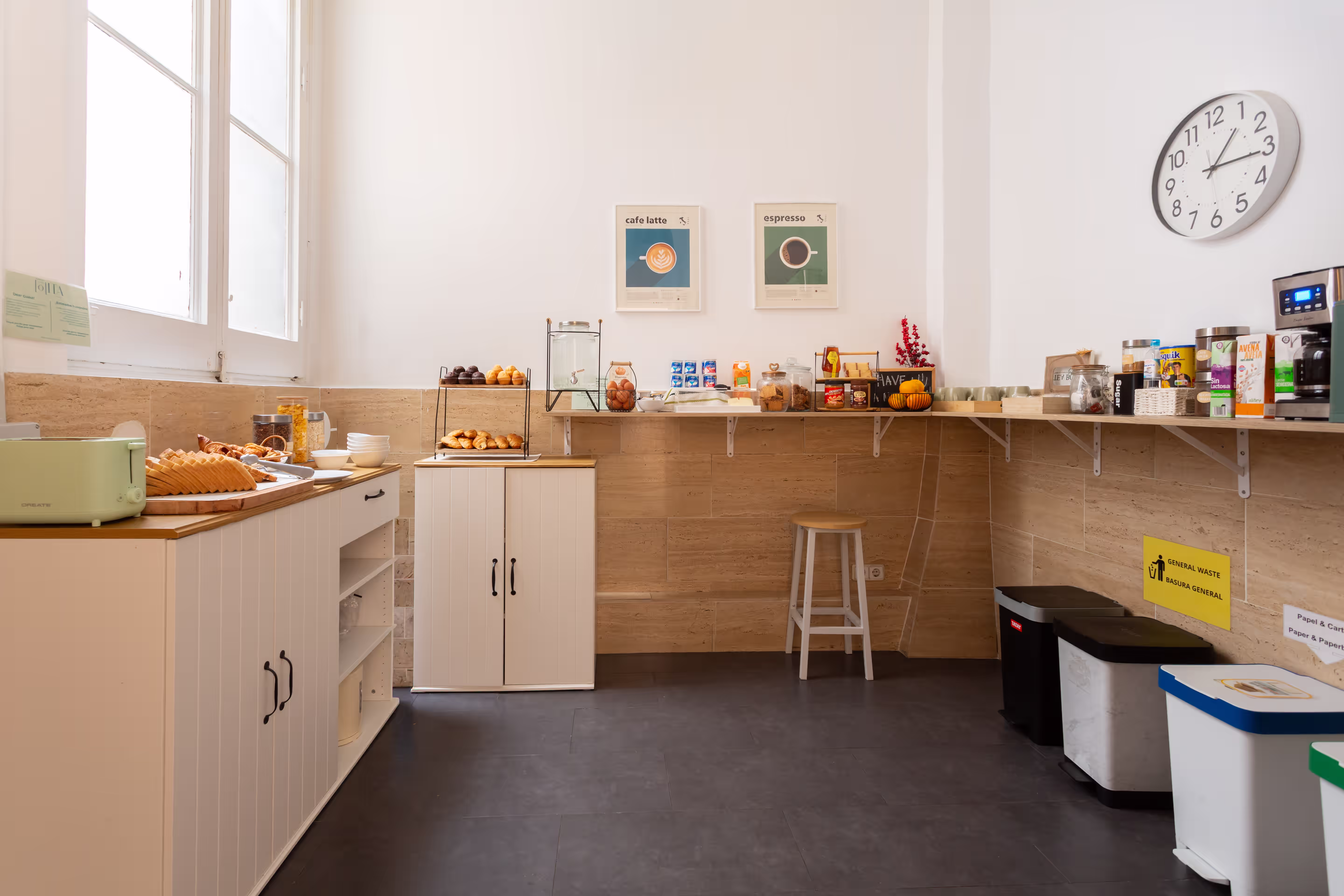 Bright kitchen corner with pastries, cereal, drinks, coffee machine, and a clock on the wall.