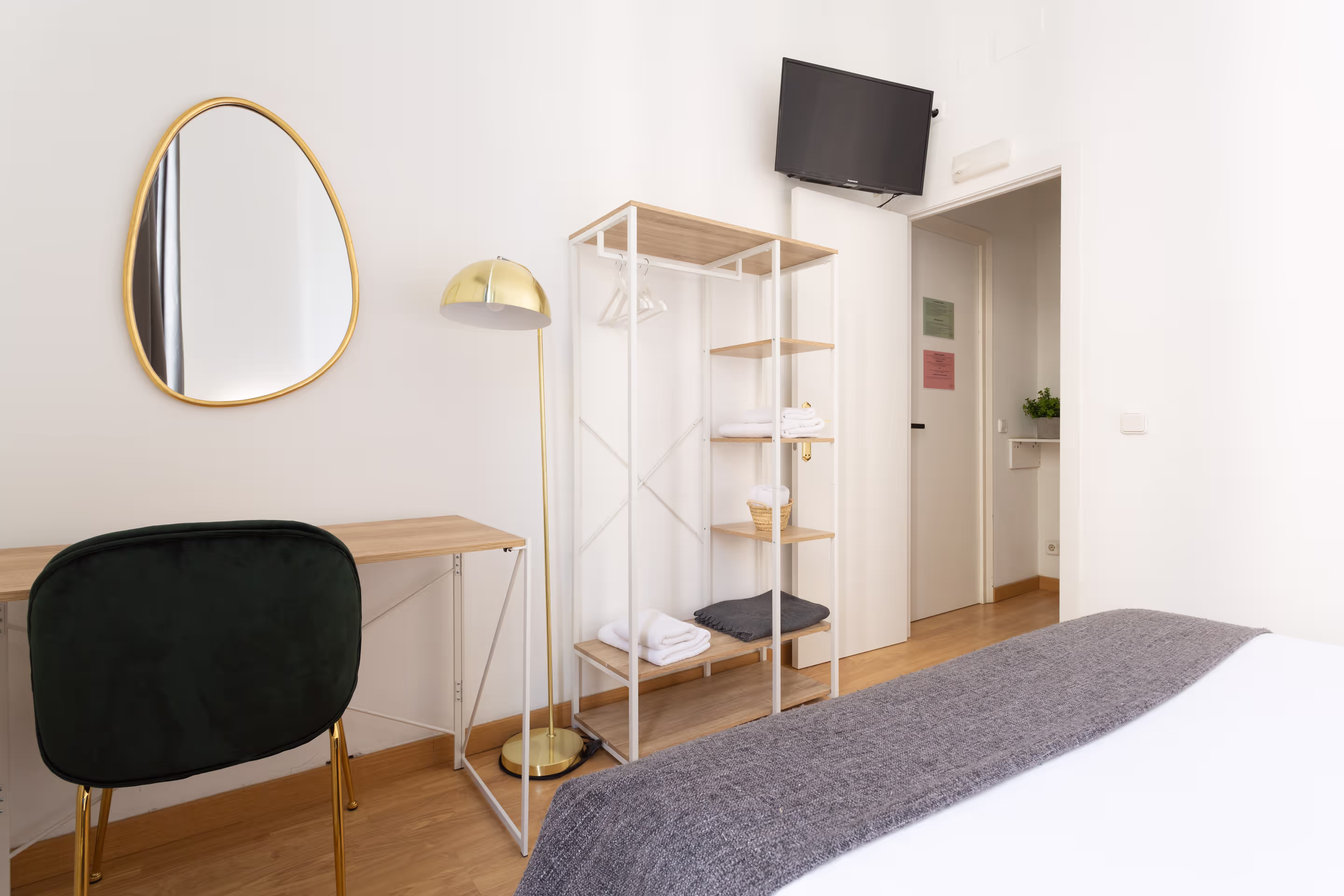 Minimalist bedroom corner with a black chair, wooden desk, gold floor lamp, shelves, and wall-mounted TV.