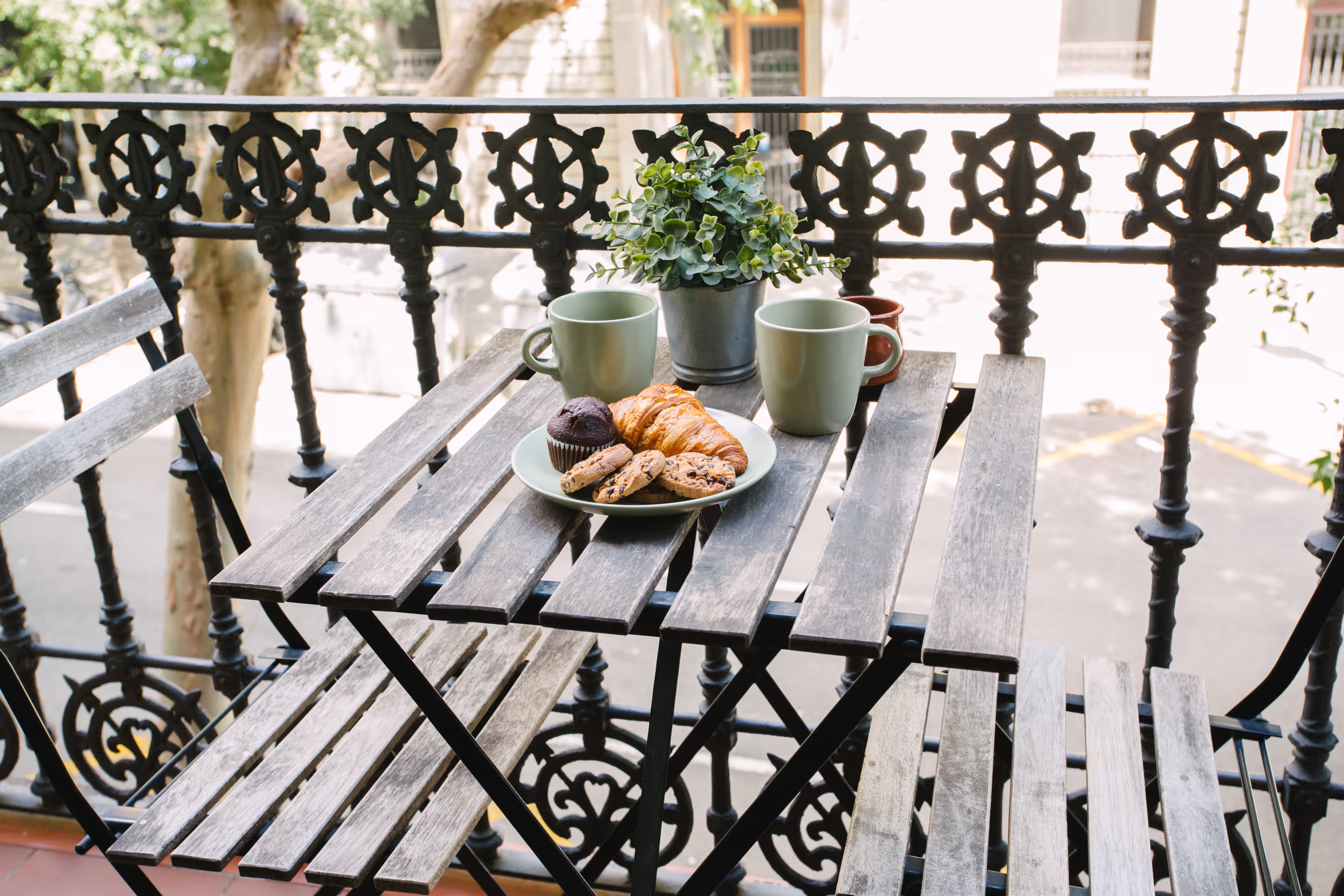 Wooden balcony table with two green mugs, plate of pastries and cookies, and a small potted plant.