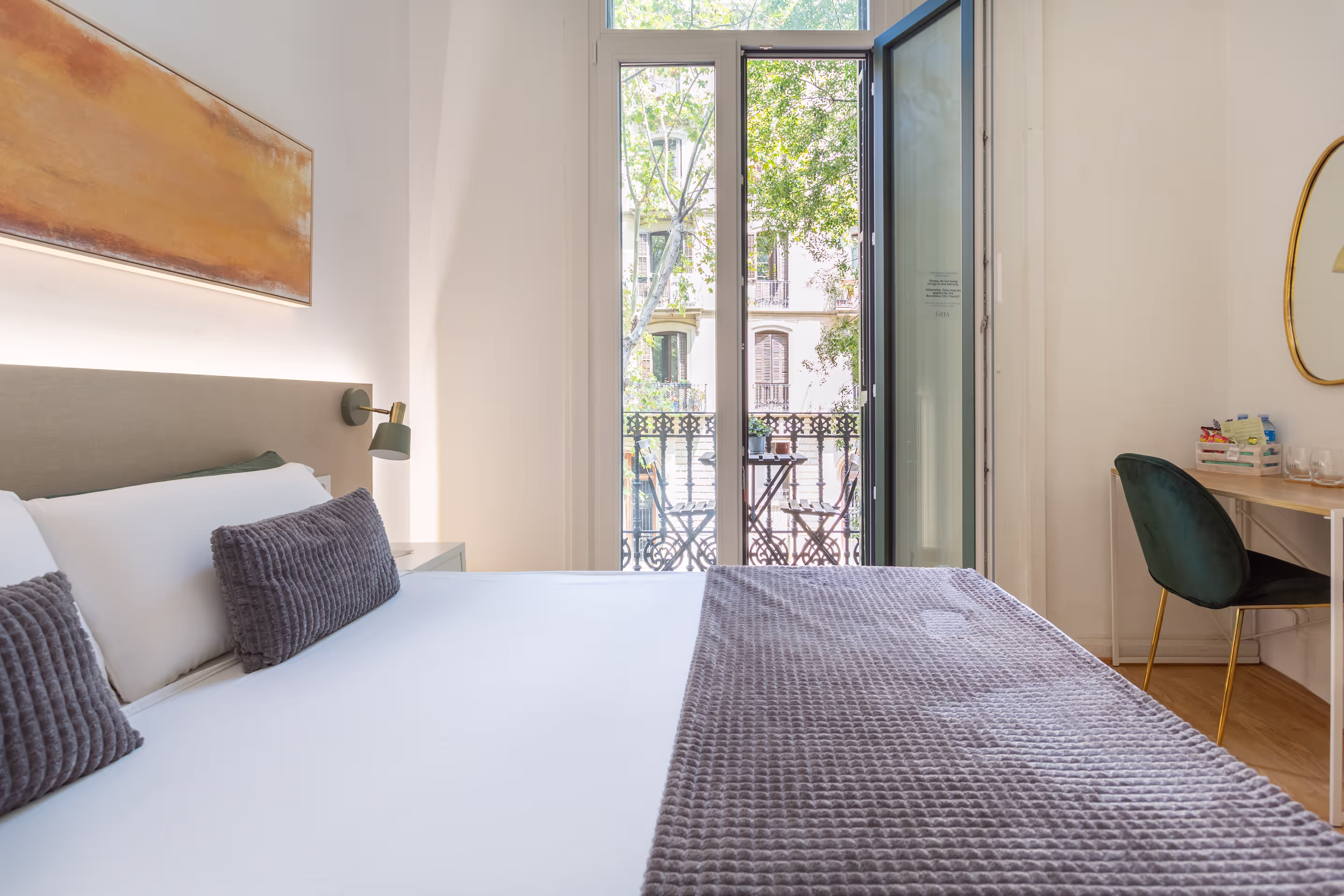 Bright bedroom with a large bed, plush pillows, open balcony door, and a desk with chair and mirror.