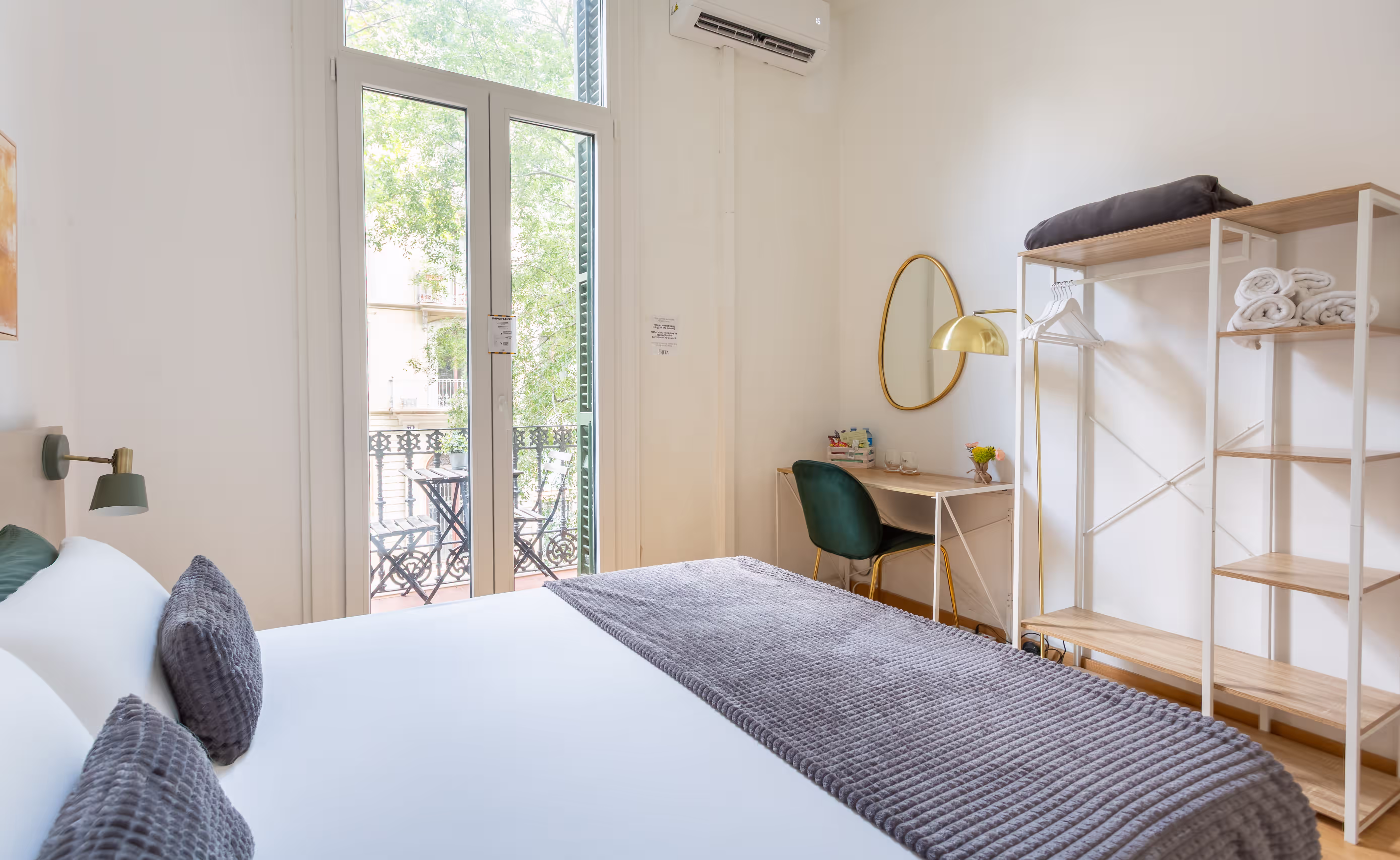 Bright bedroom with a large bed, gray blanket, balcony with chairs, a desk, and open shelving with towels.