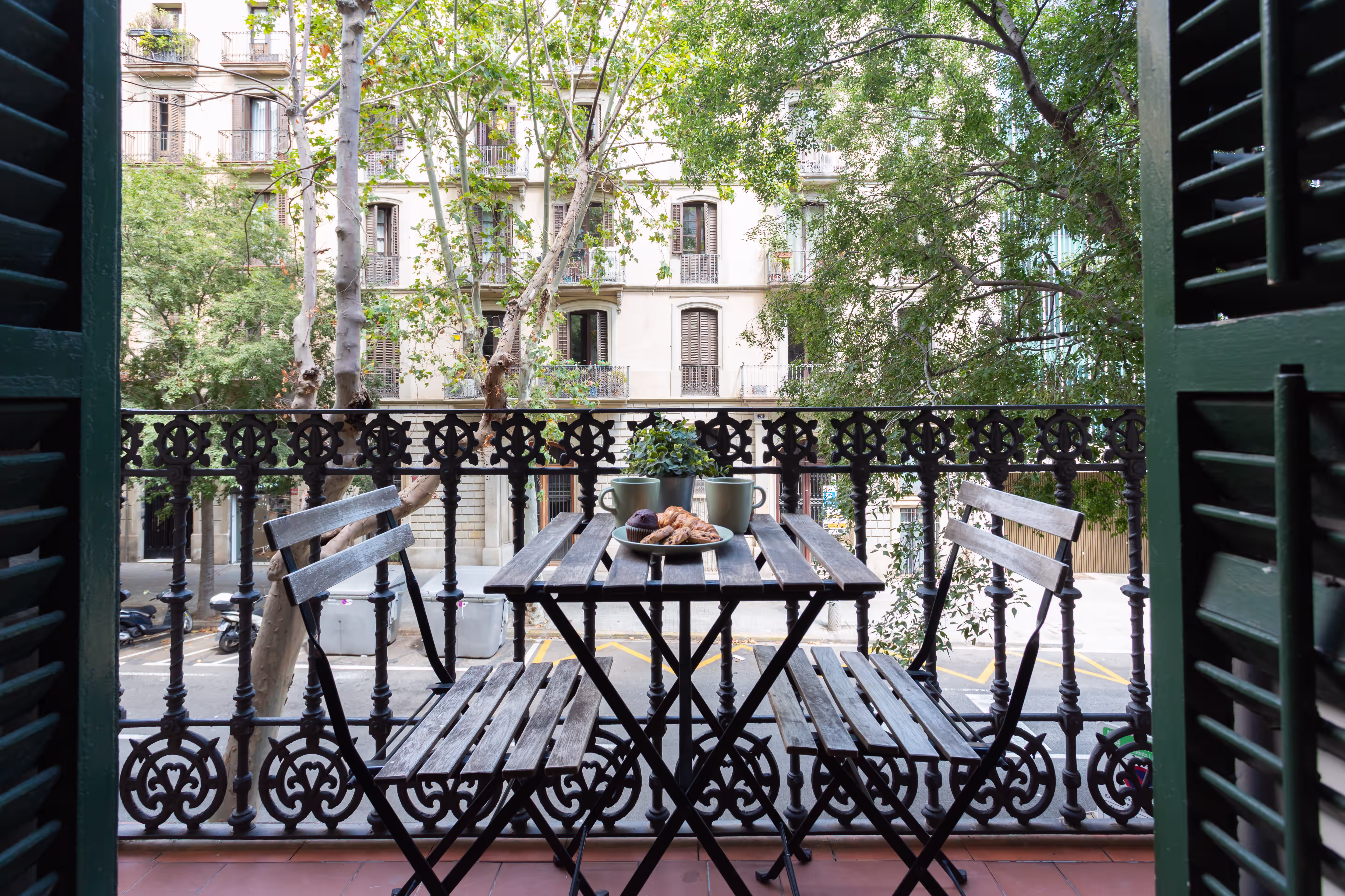 Small balcony with a wooden table and chairs, two mugs, pastries, and a plant overlooking a street.