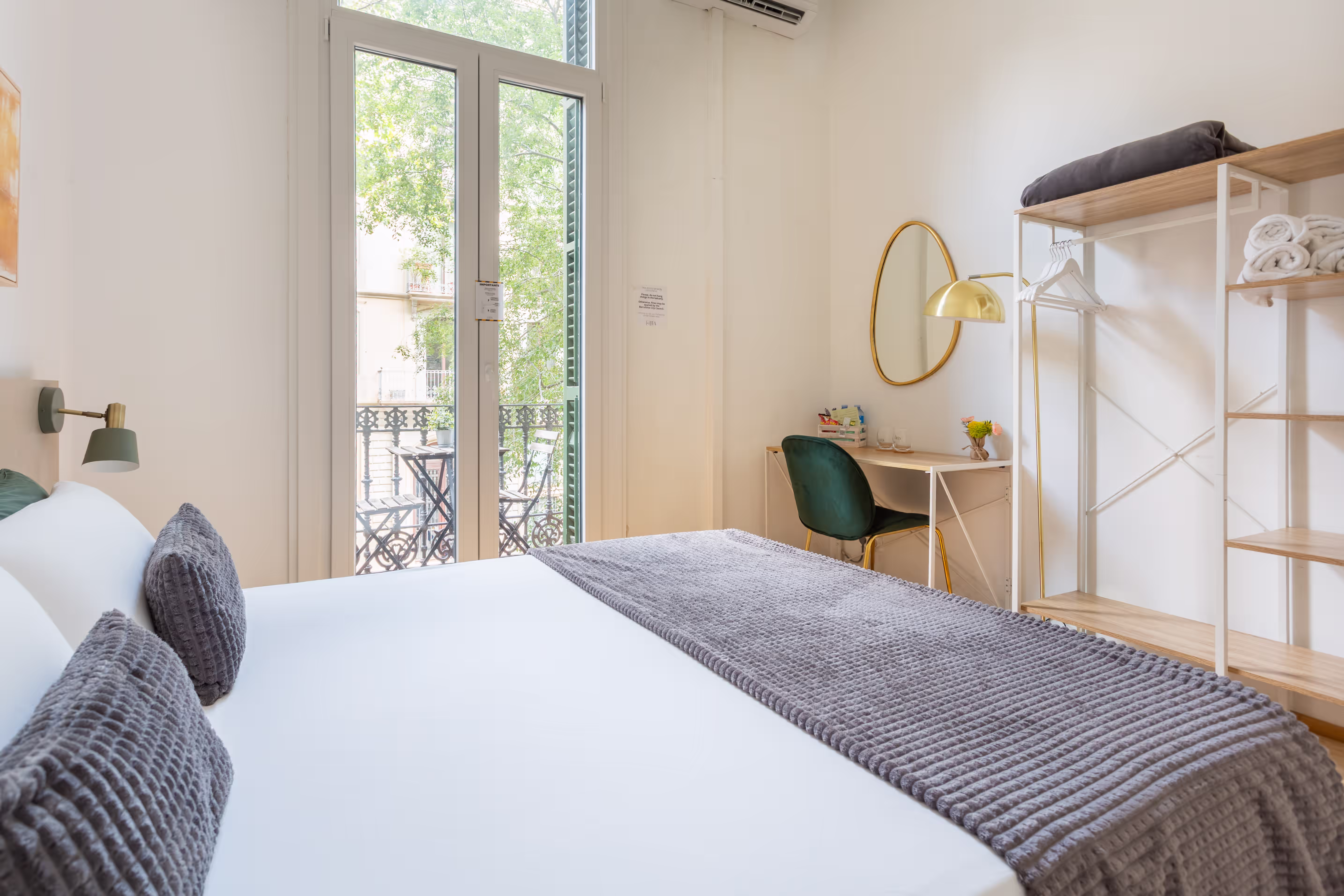 Bright bedroom with white bed, gray pillows, balcony door, desk, green chair, and wooden shelves.
