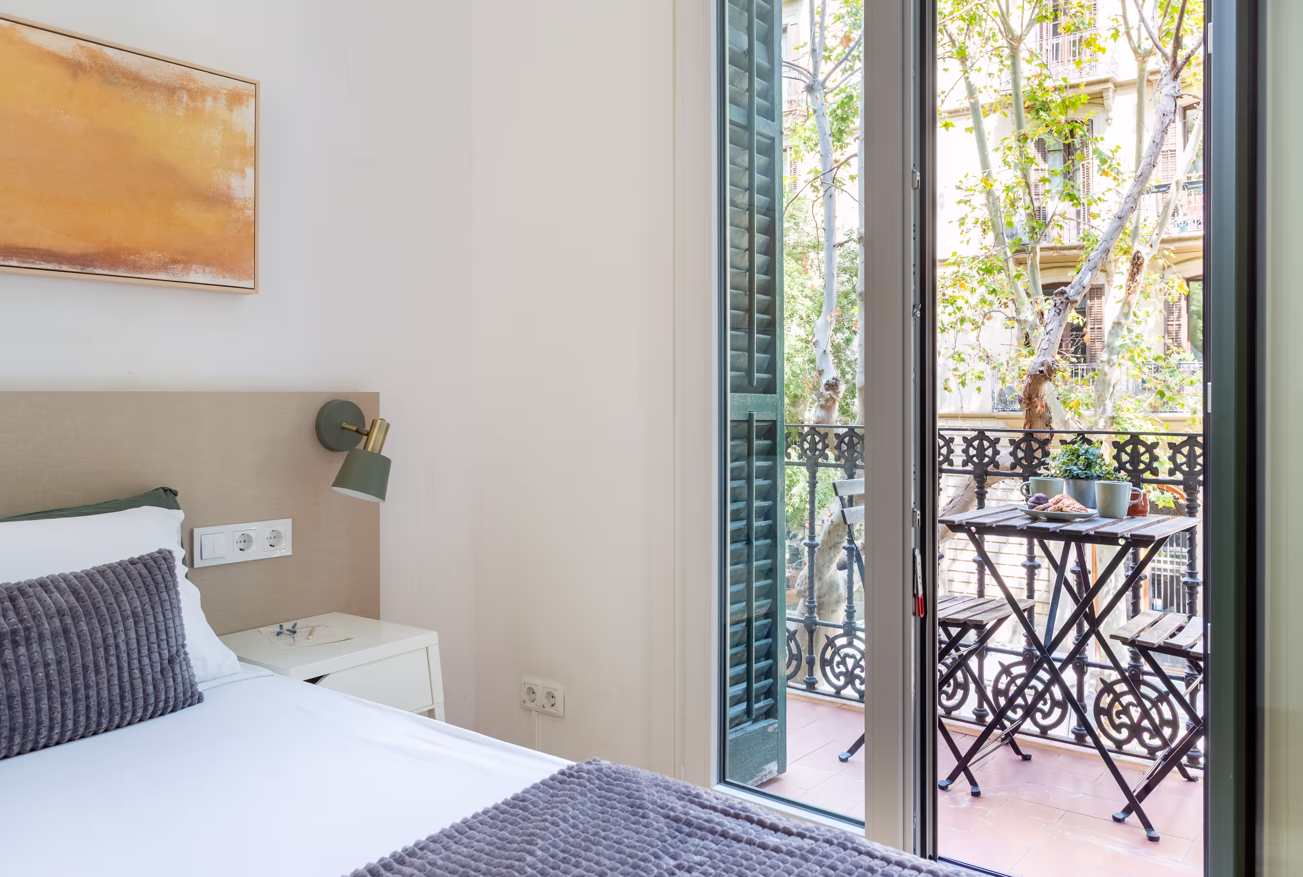 Bright bedroom with white and gray bedding next to a balcony with a table, chairs, and plants.