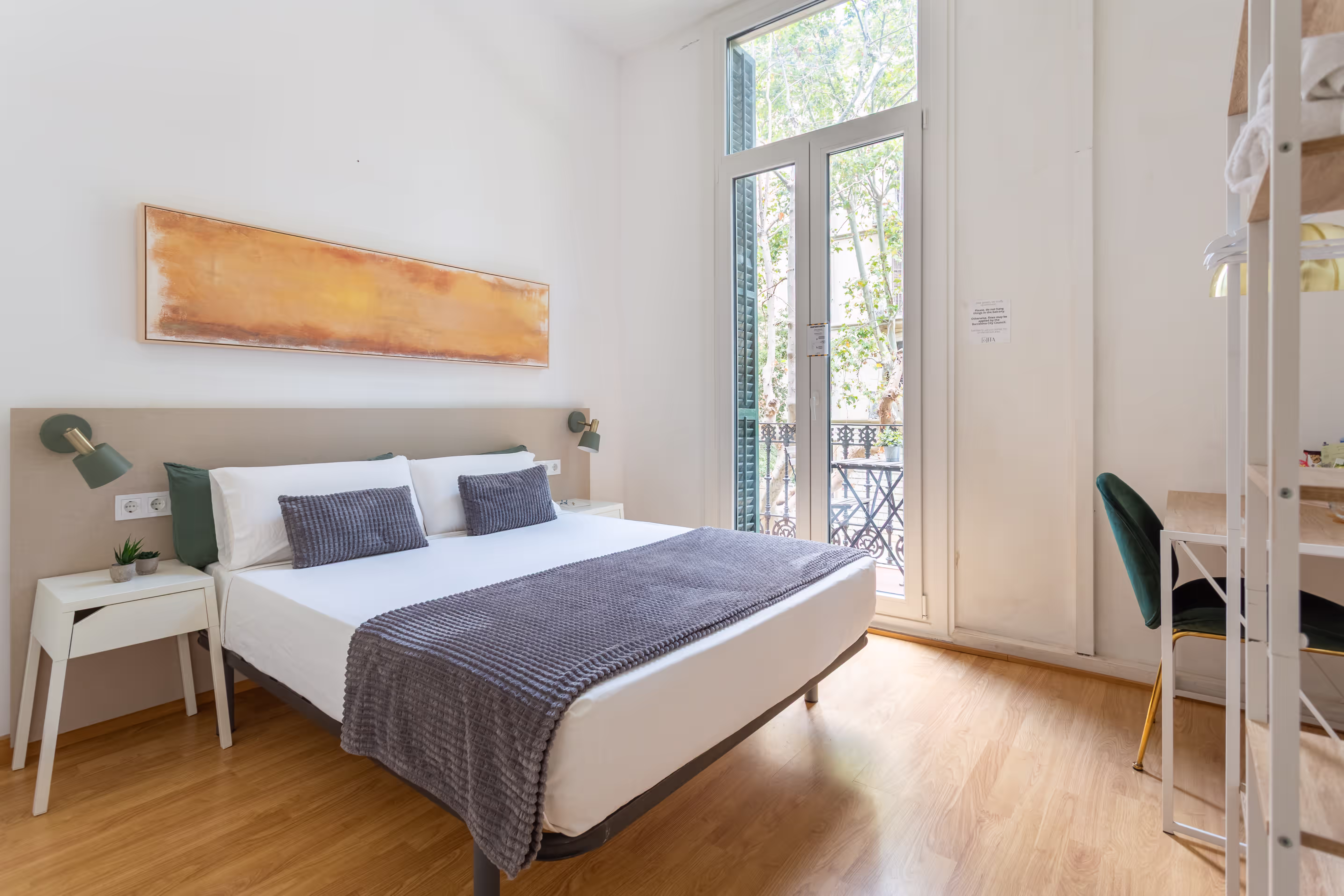 Bright bedroom with large bed, gray pillows and blanket, side tables, and a tall window with balcony.