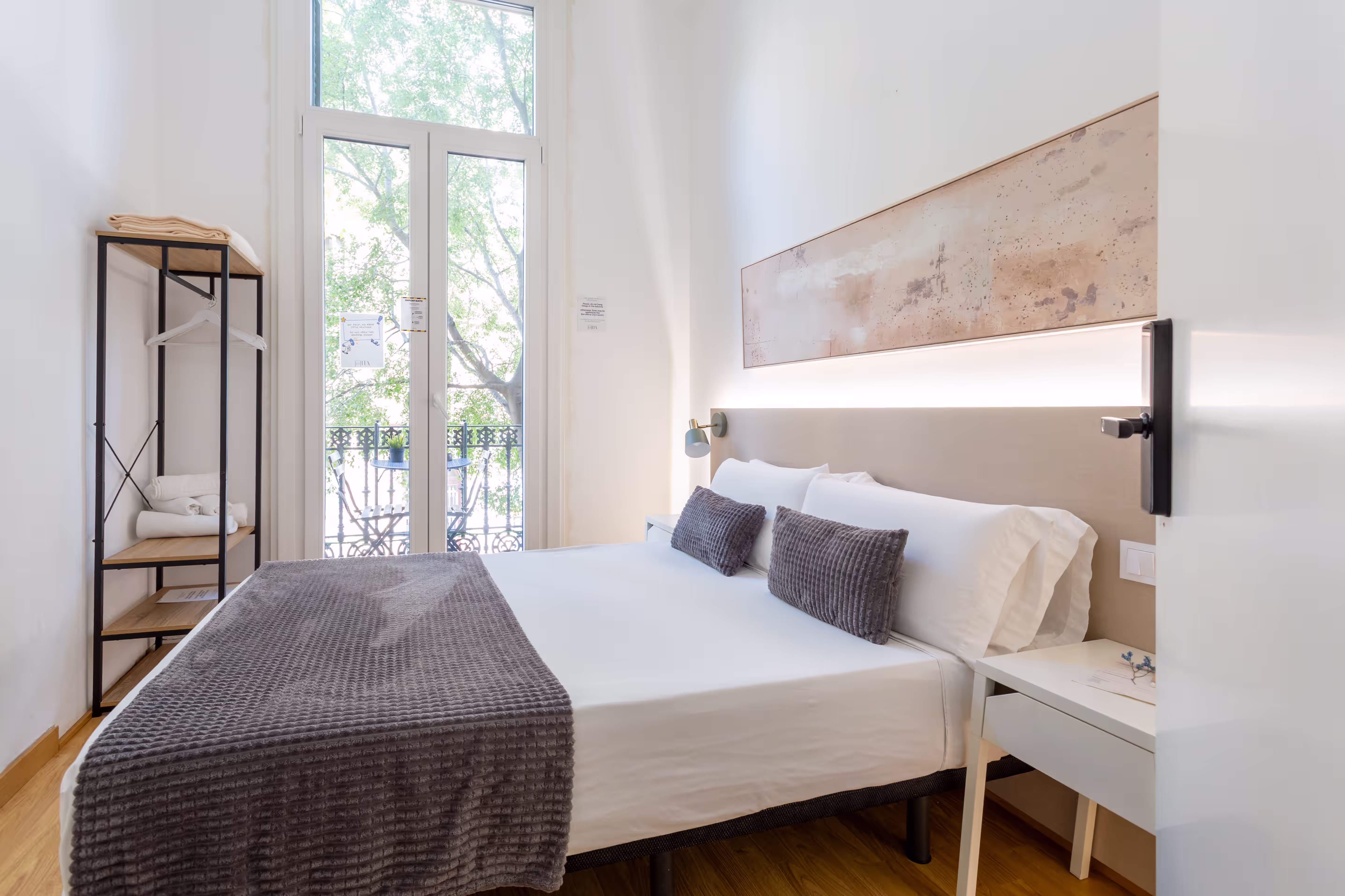 Bright bedroom with a double bed, gray throw and pillows, minimalist rack, and large window with balcony view.