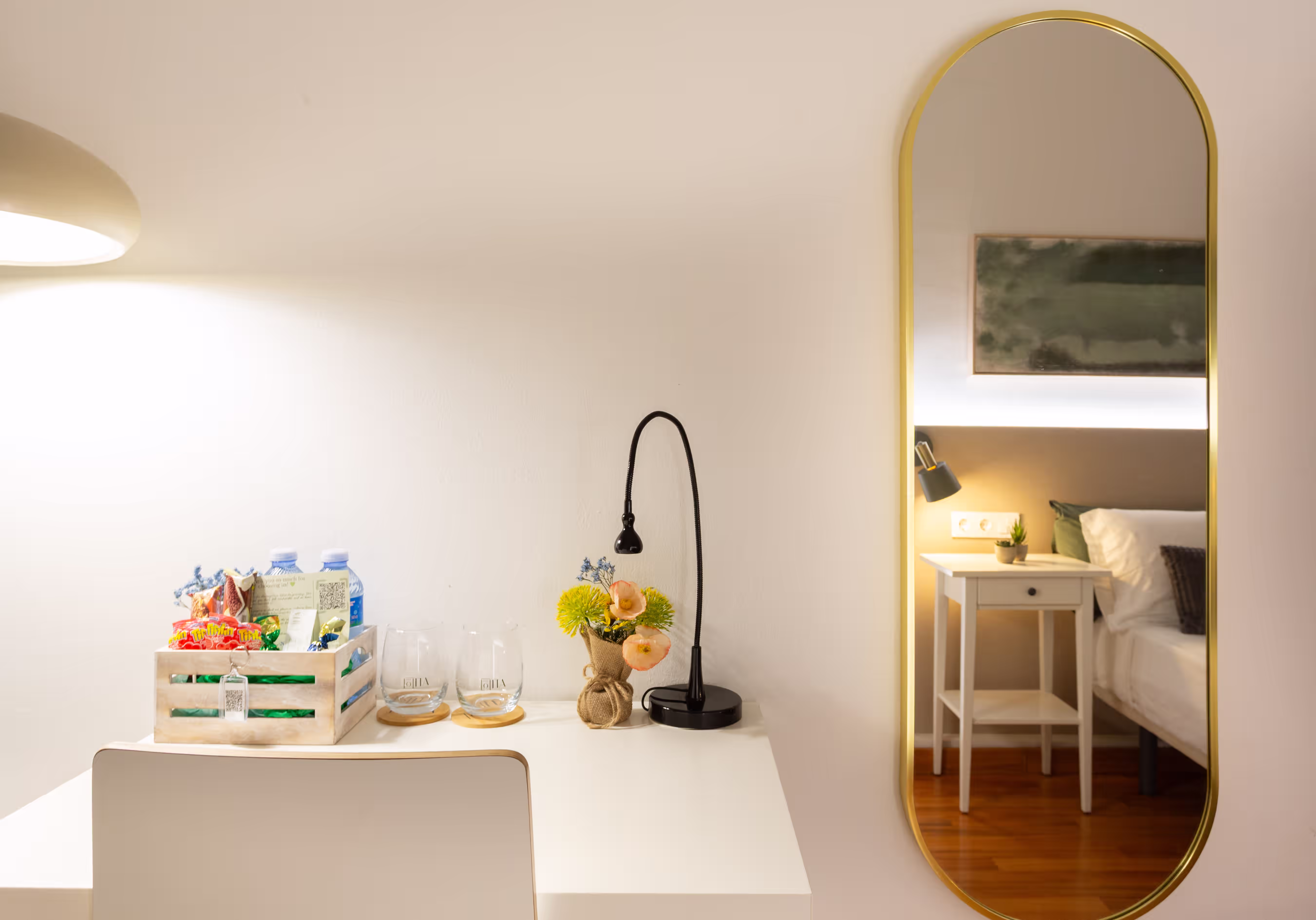 White desk with lamp, flowers, snacks, two glasses, and a gold-framed mirror reflecting a bedside table and bed.