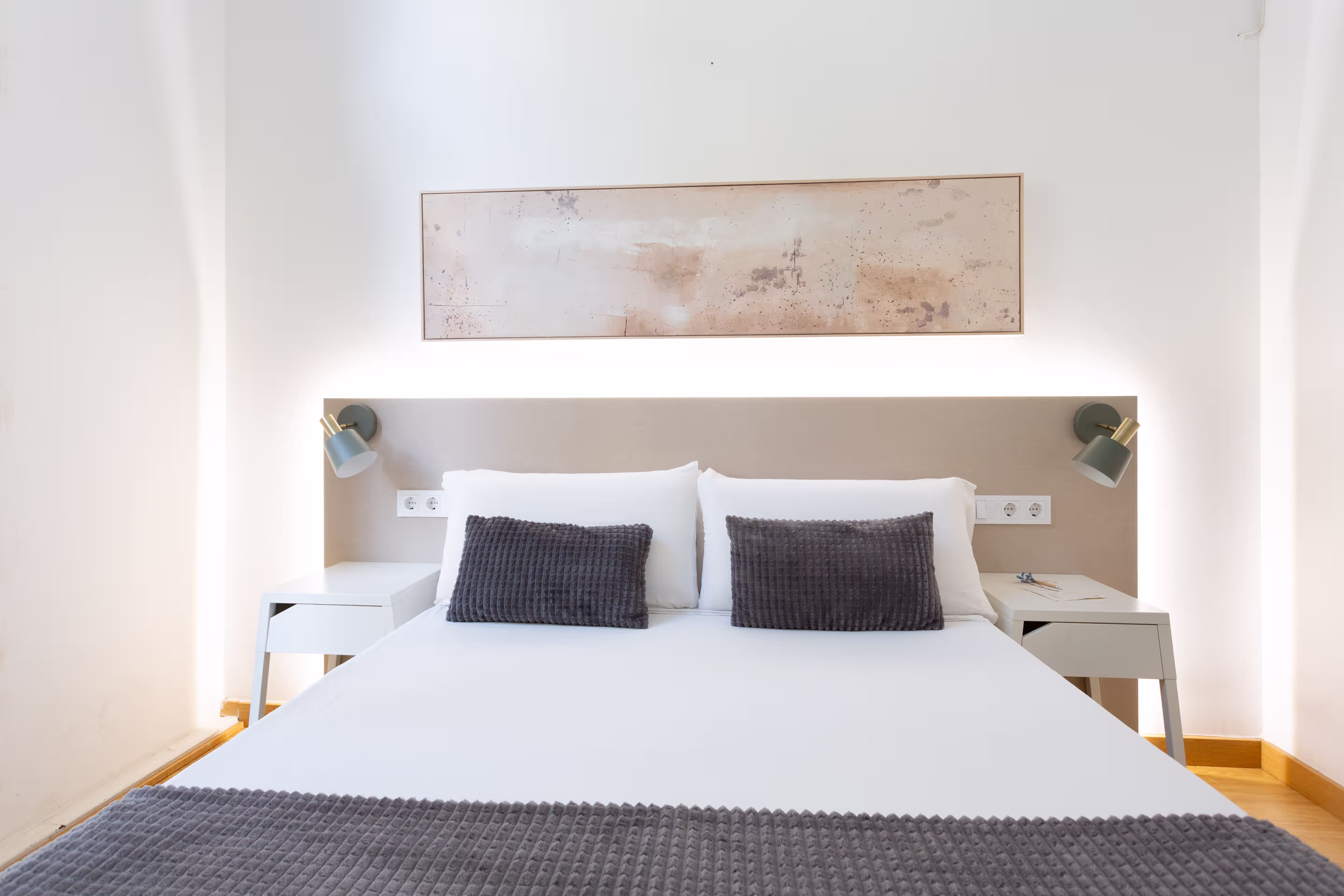 Minimalist bedroom with white bed, gray textured pillows and throw, two white nightstands, wall lamps, and abstract art.