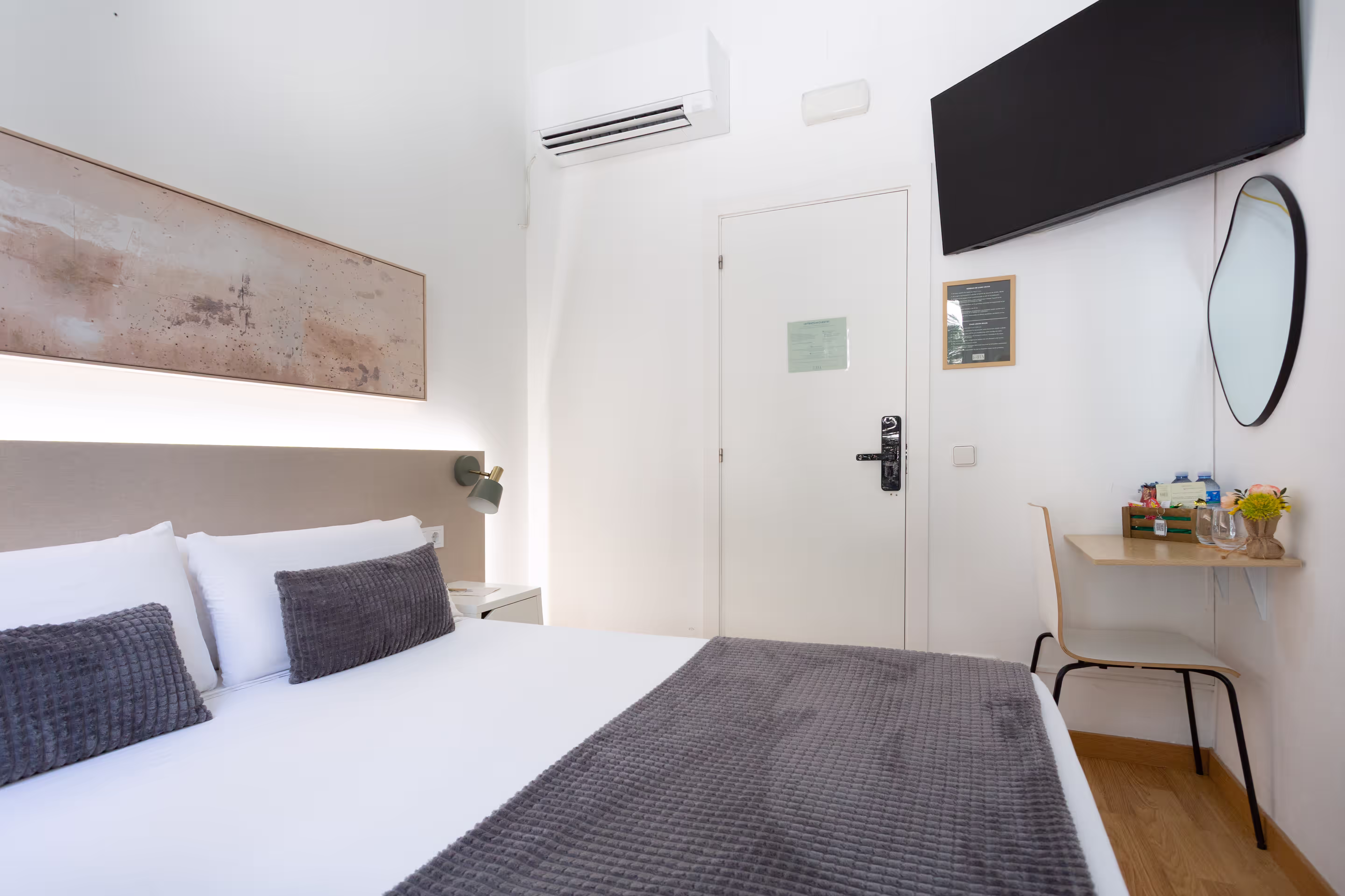Modern hotel room with a large bed, gray bedding, wall-mounted TV, desk, and air conditioner.