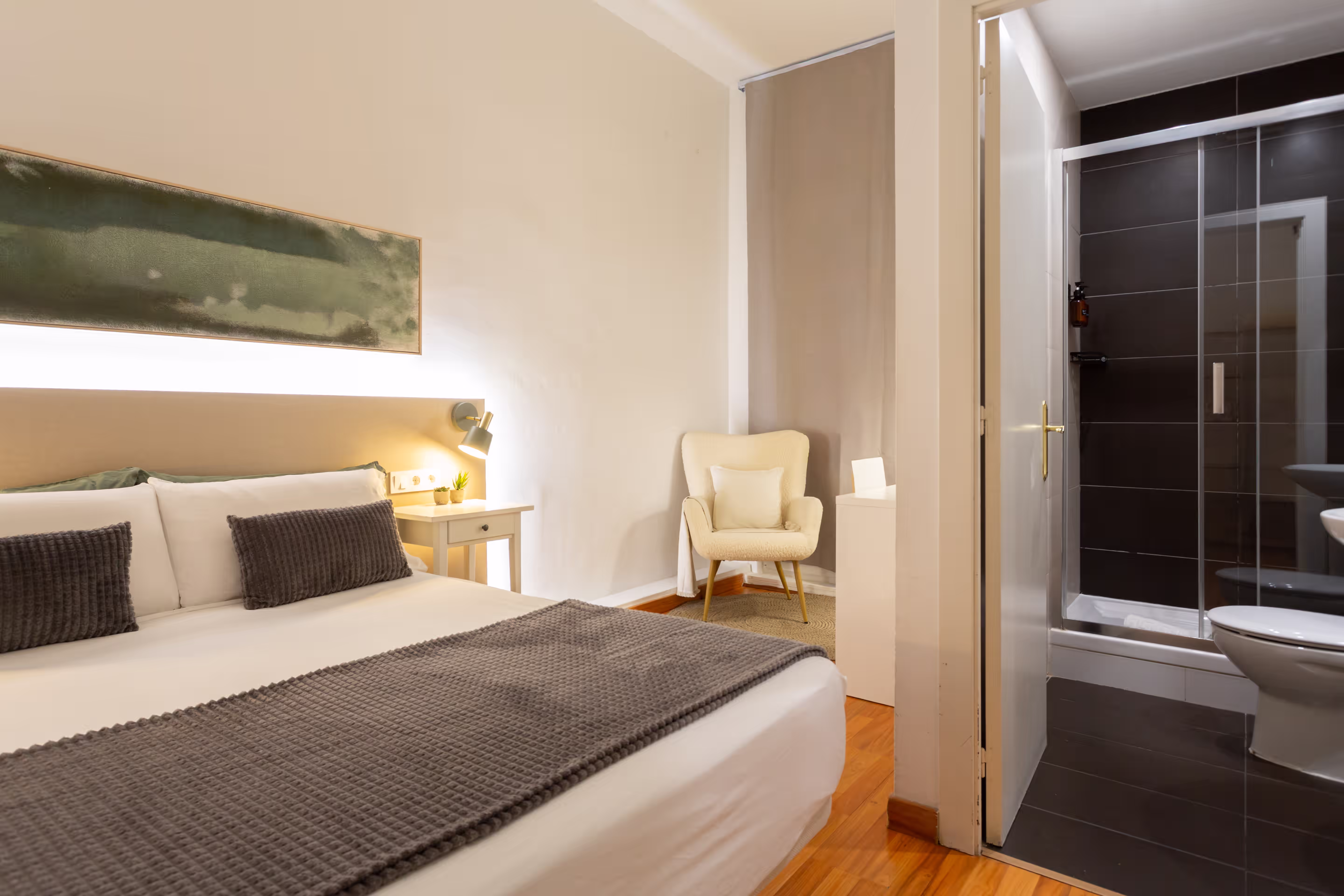 Modern bedroom with a white bed, gray pillows and blanket, armchair, and an adjacent bathroom.