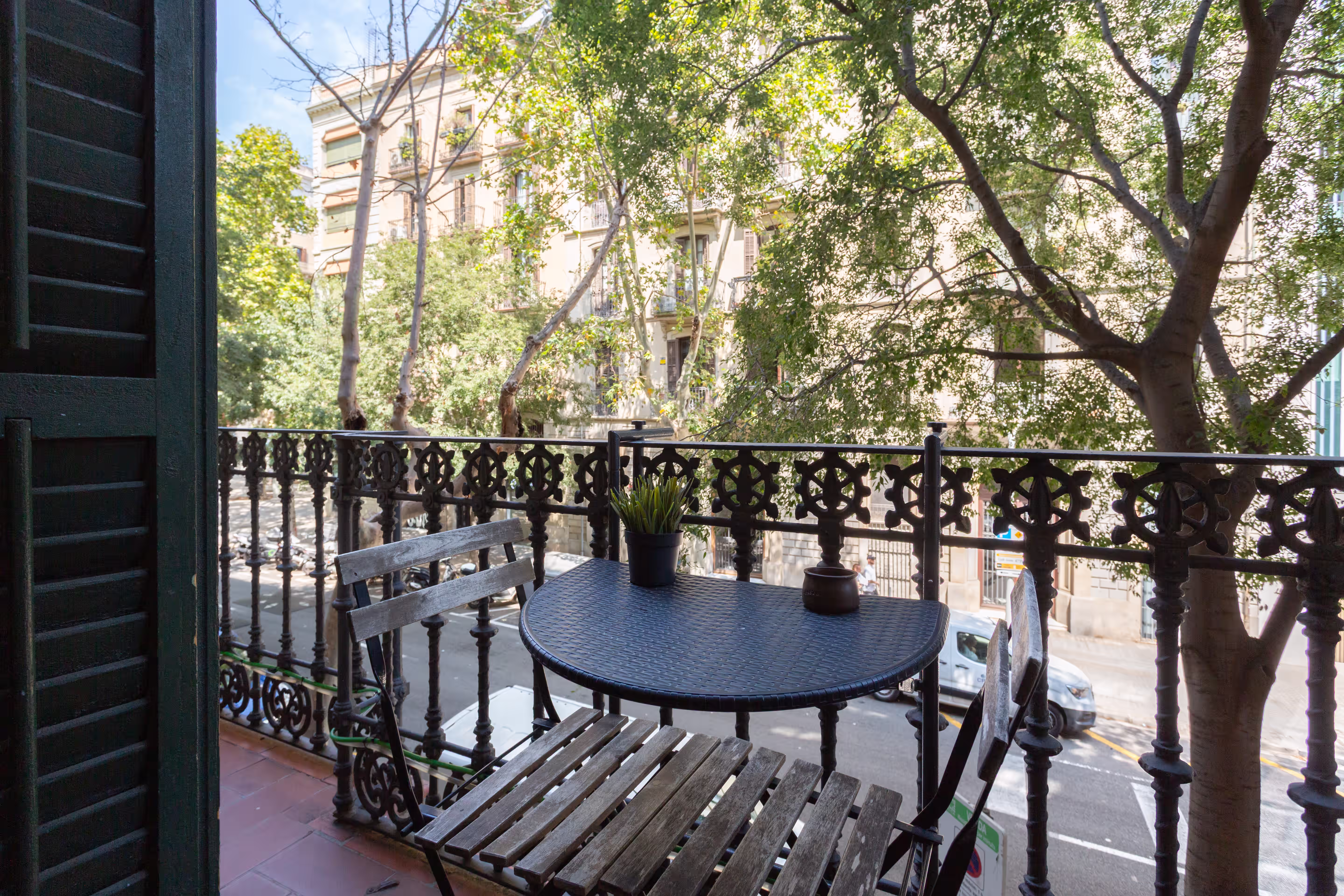 Small balcony with two wooden chairs and a round table overlooking a tree-lined street.