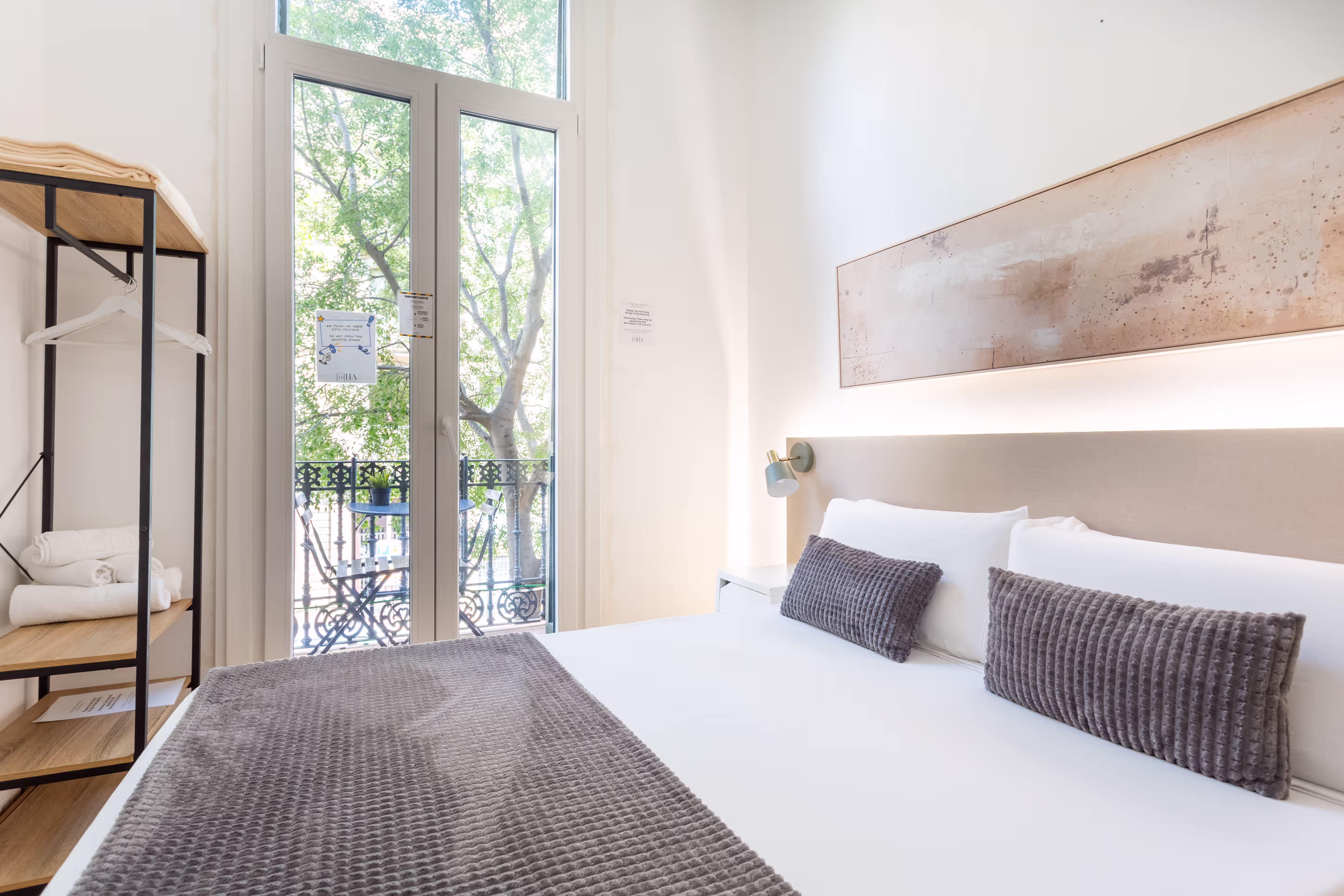 Bright hotel room with king bed, gray textured pillows and throw, balcony with black chairs and table.